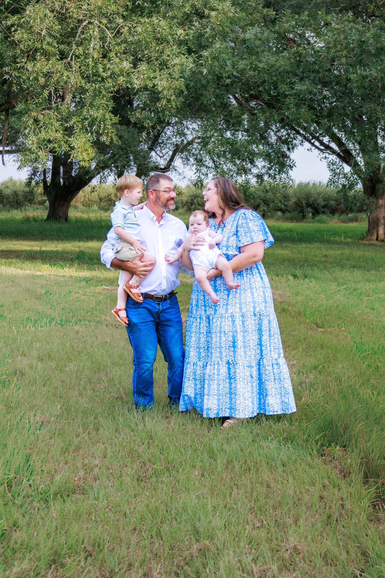Family of four posing outdoors in green field, parents holding two young children, woman in blue floral dress.