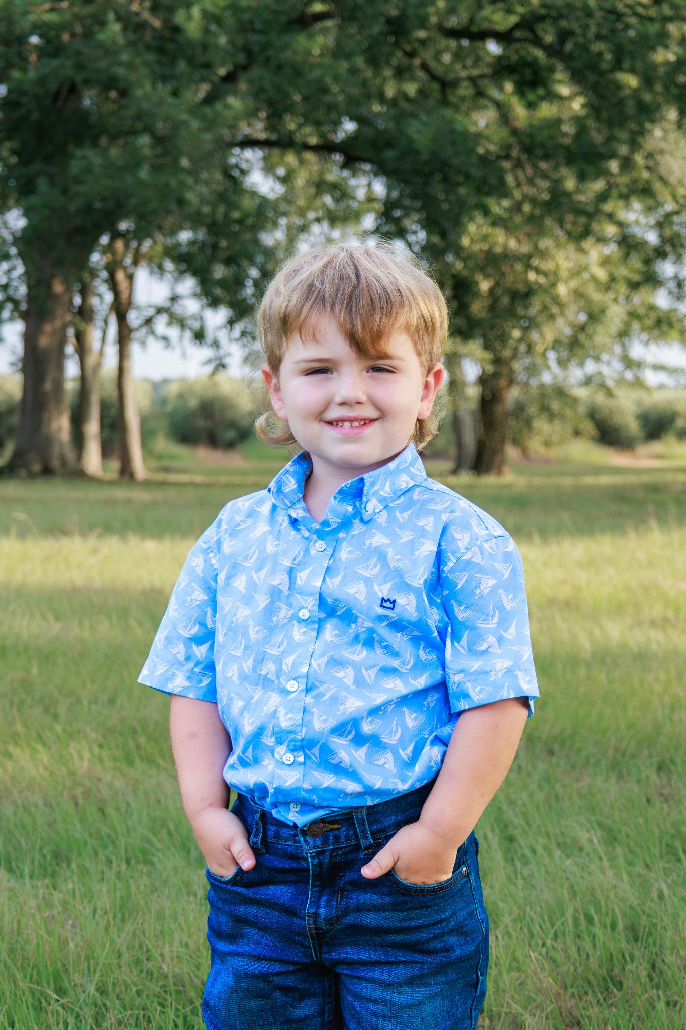 Smiling young boy in a blue patterned shirt and jeans standing in a green field with trees.