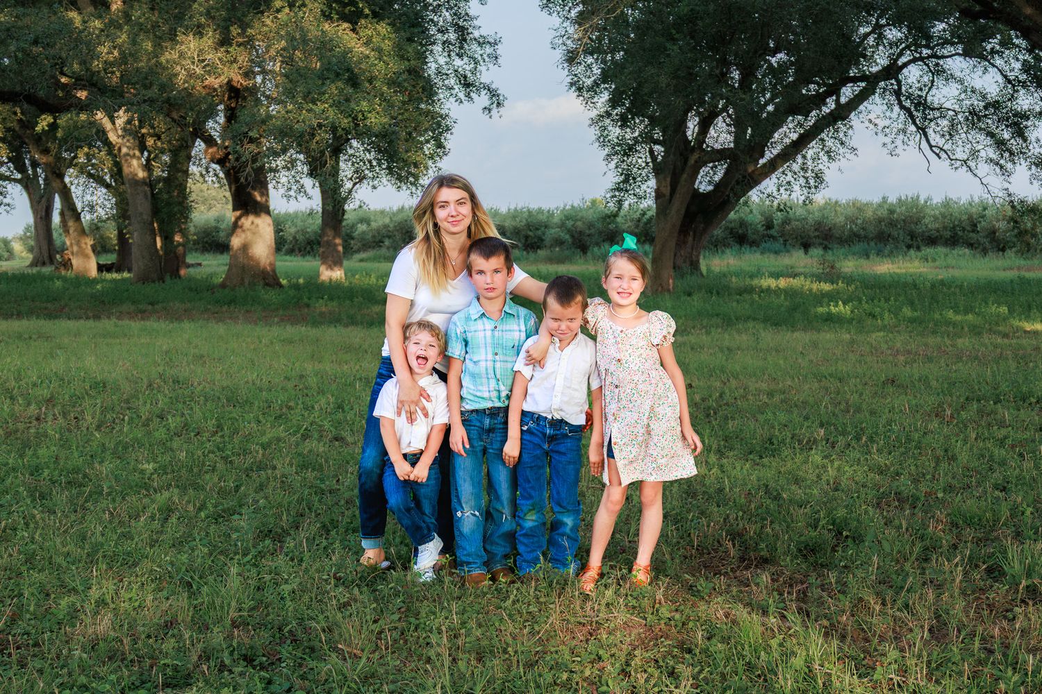 A woman poses outdoors on green grass with four children under large trees in a warm family portrait.