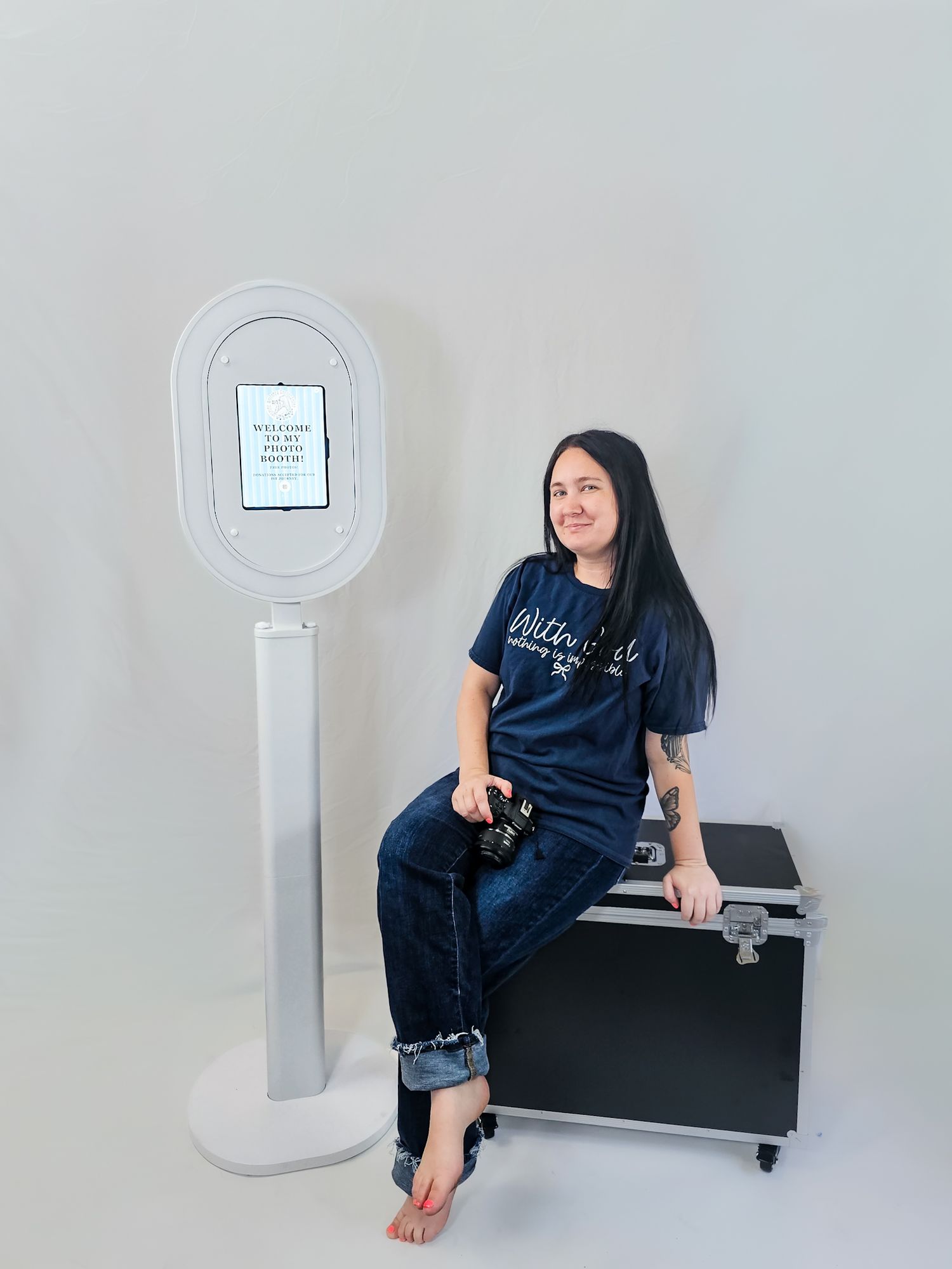 Woman in dark t-shirt sits on a travel case next to a modern photo booth kiosk on a white background.