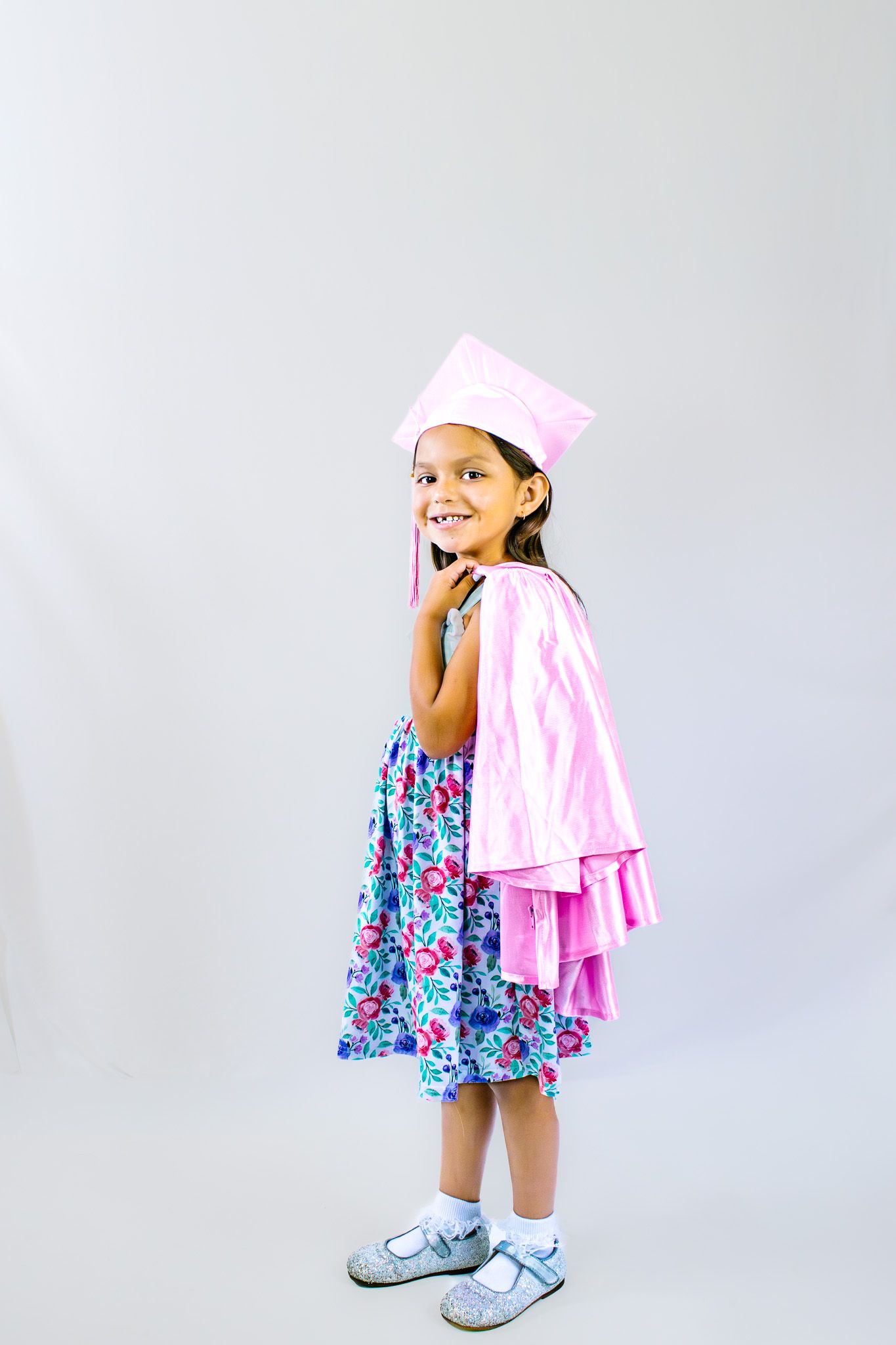 Smiling young girl wearing a pink graduation cap and gown over a floral dress, posing proudly for a photo.