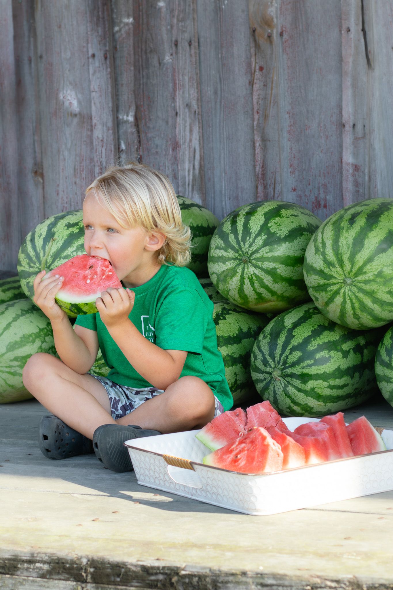Young blond boy in green shirt eating watermelon slice beside sliced watermelon tray with whole melons stacked behind him.