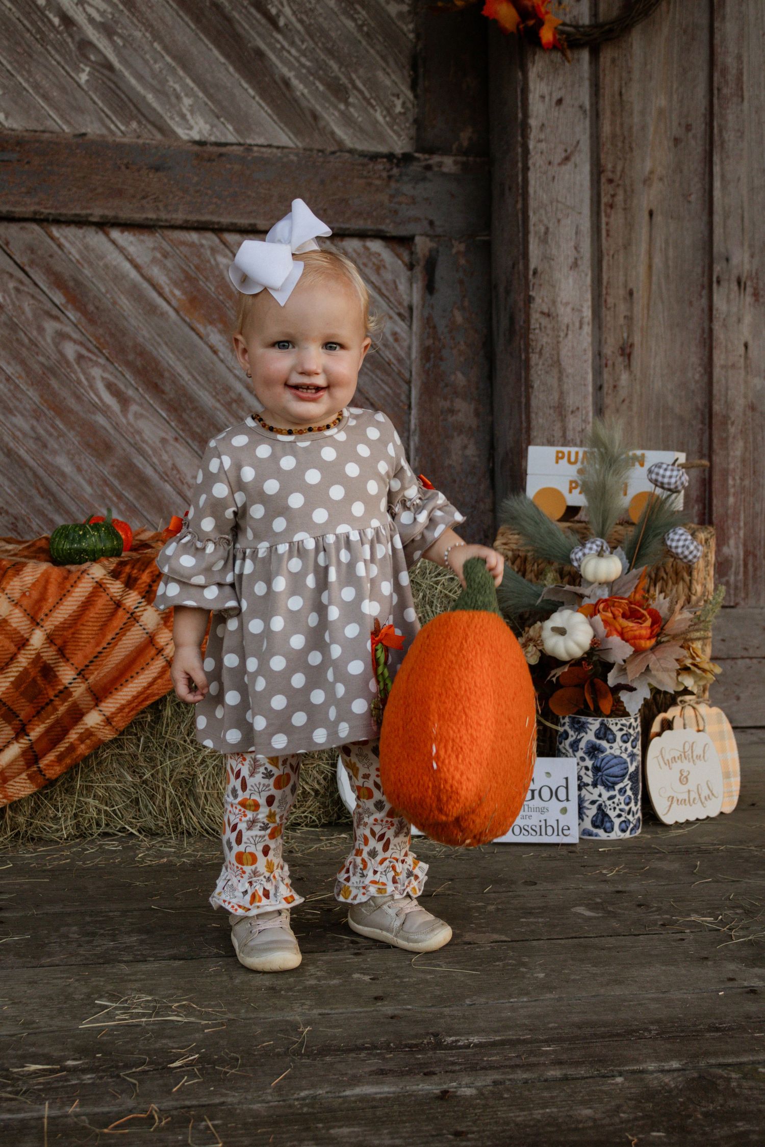 Smiling toddler girl in polka dot dress holding large orange pumpkin in fall barn setting.