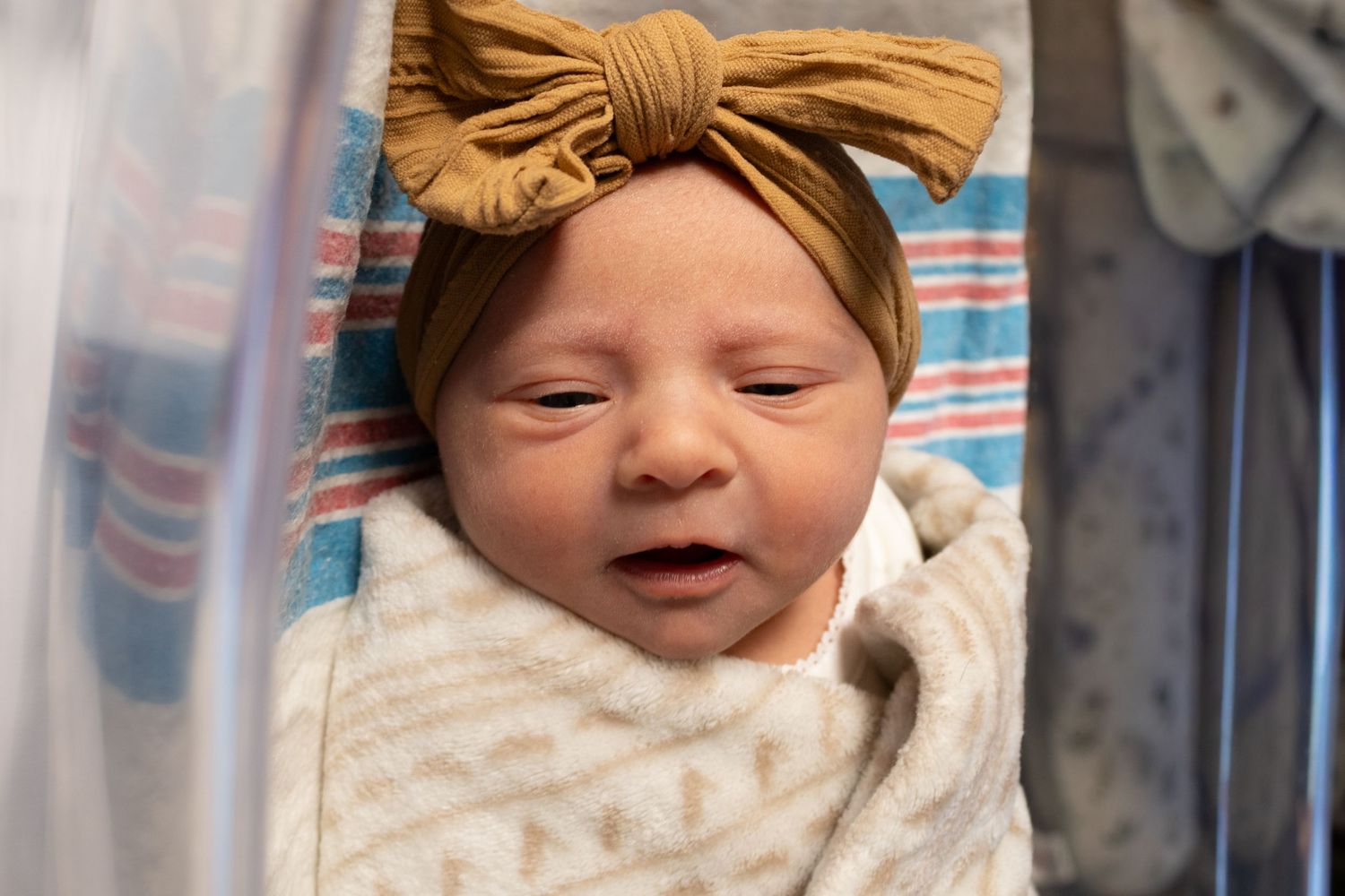 Newborn baby girl swaddled in striped hospital blanket wearing mustard yellow bow headband in bassinet.
