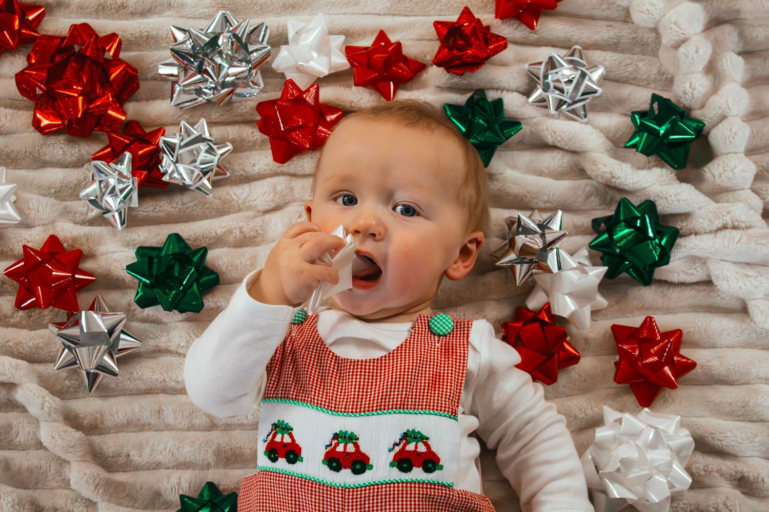 Baby in Christmas romper surrounded by colorful holiday bows on cozy blanket.