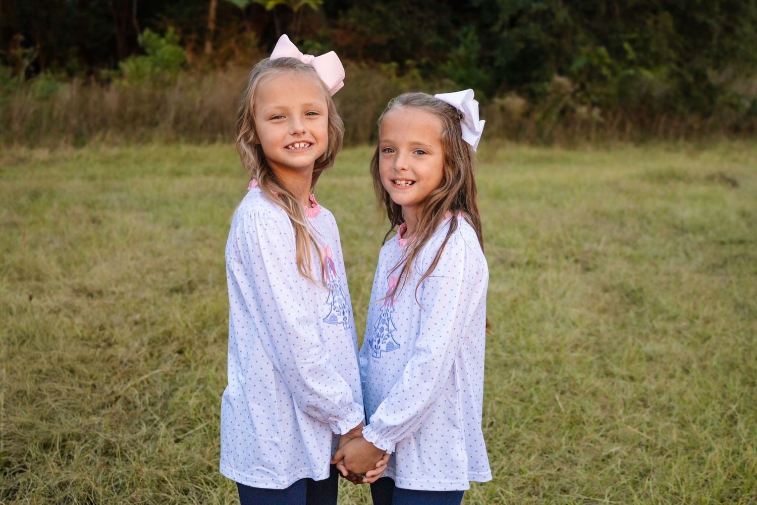 Two young girls in matching white dresses with pink bows holding hands outdoors.