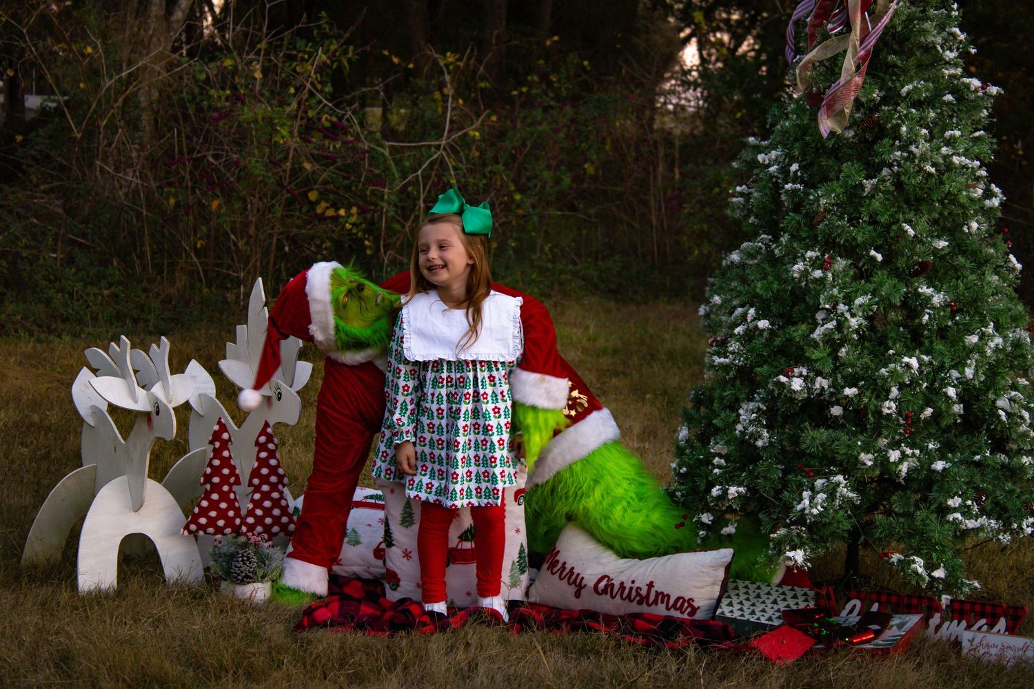 Young girl laughing with Grinch character beside Christmas tree and white reindeer decorations.