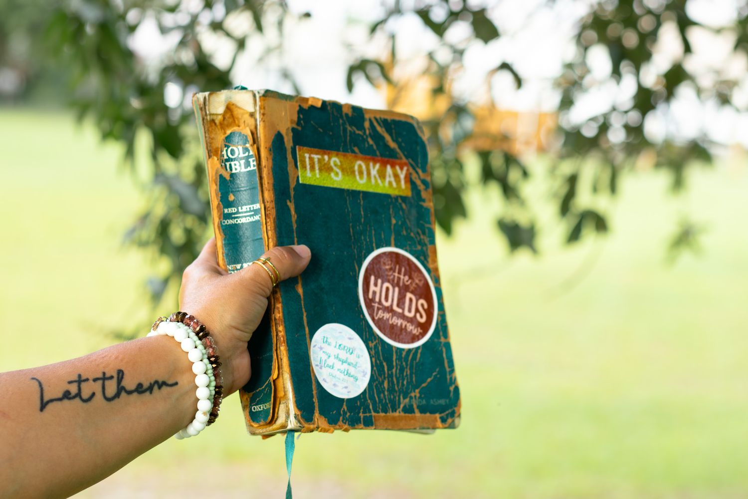 Hand holding a vintage teal book titled It's Okay with a let them tattoo visible on the wrist outdoors.