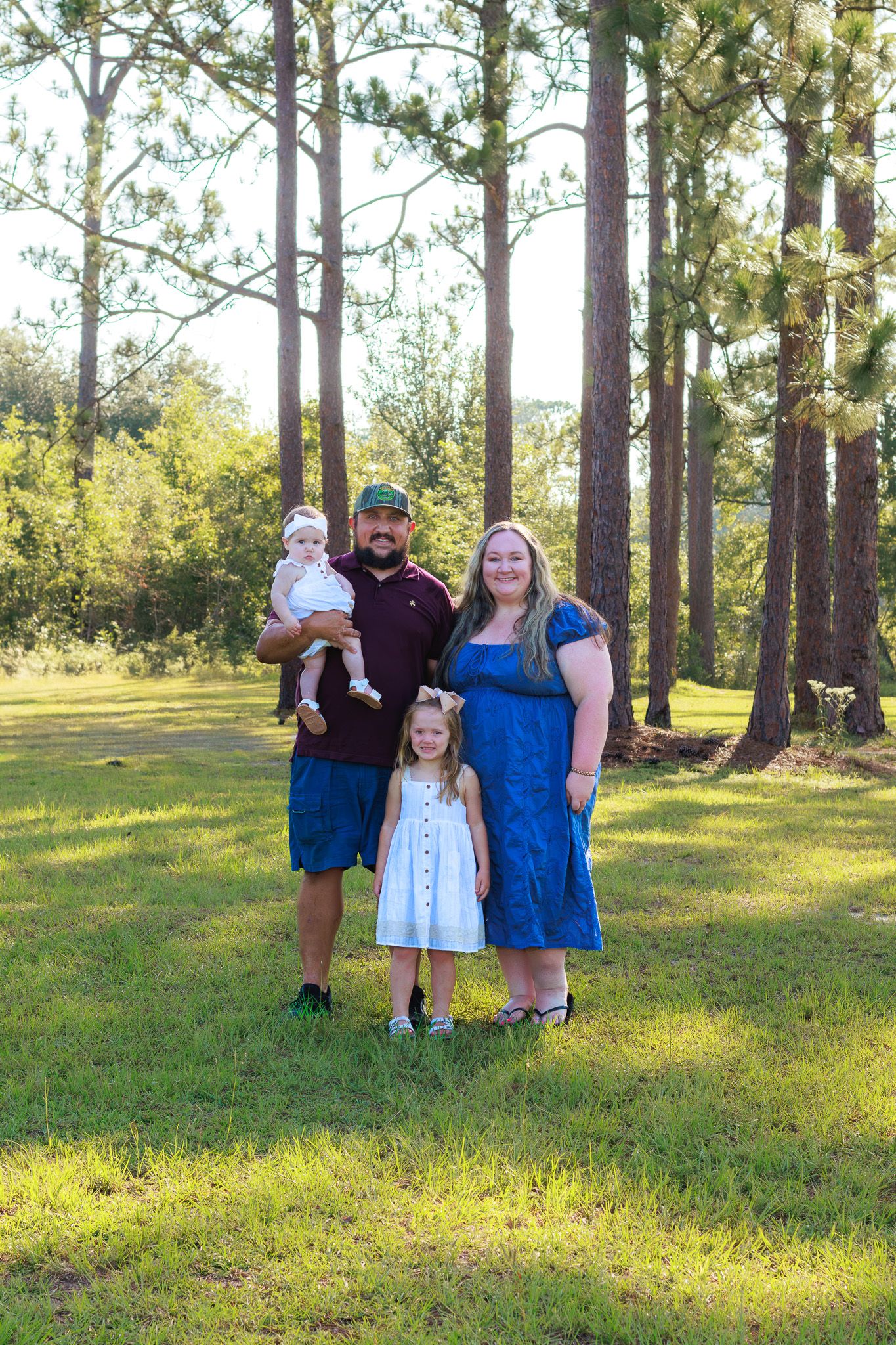 Family of four posing outdoors in a sunny pine forest, parents smiling with two young daughters in casual attire.