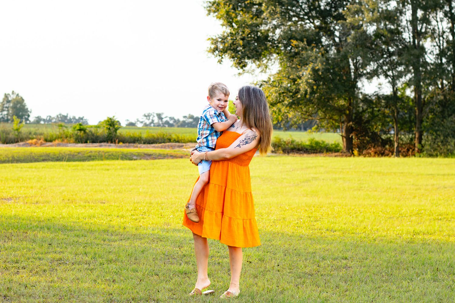 A mother in an orange dress holds her young son in a sunny green field with trees in the background.