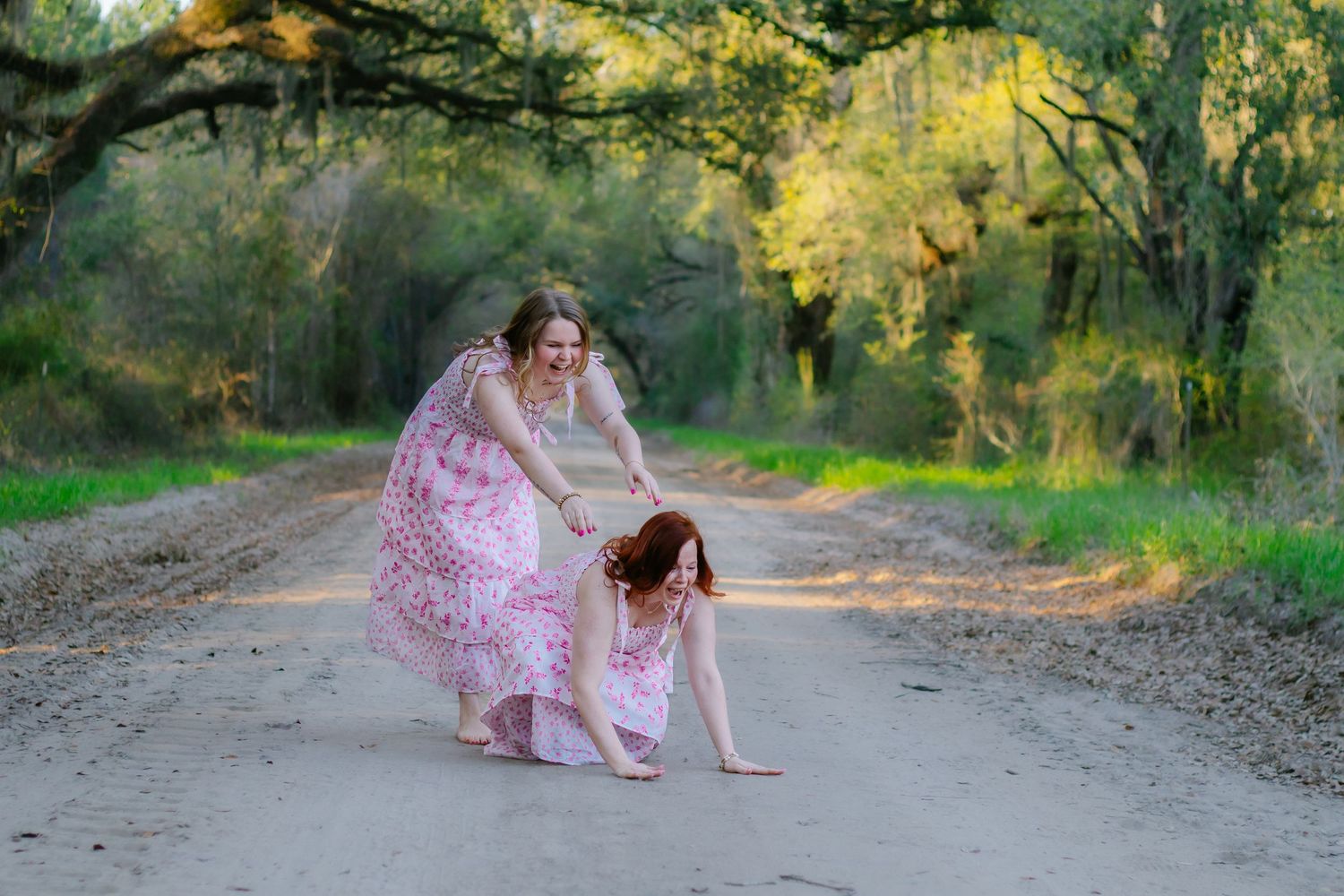 Two women in pink dresses play on a dirt road surrounded by lush green trees.