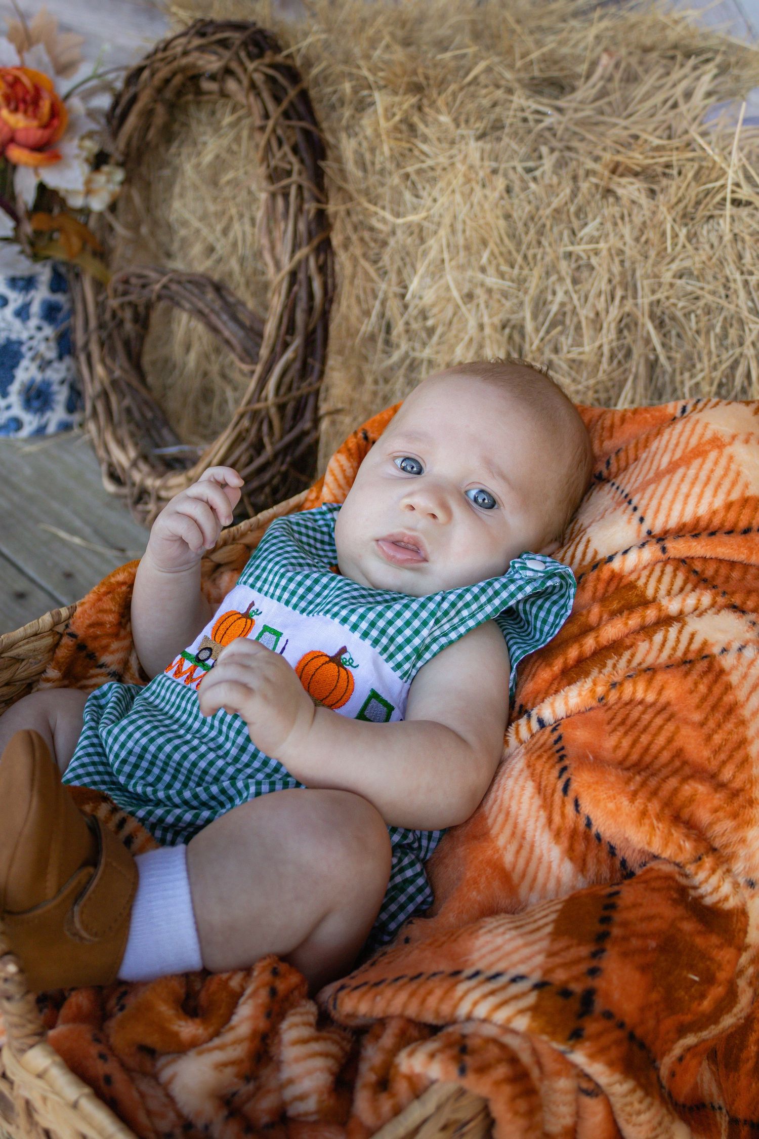 A baby in a green gingham pumpkin outfit lies in a wicker basket with fall decor.