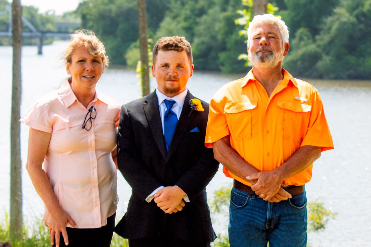 Three adults posing outdoors near a river, center man in suit with boutonniere flanked by two others.