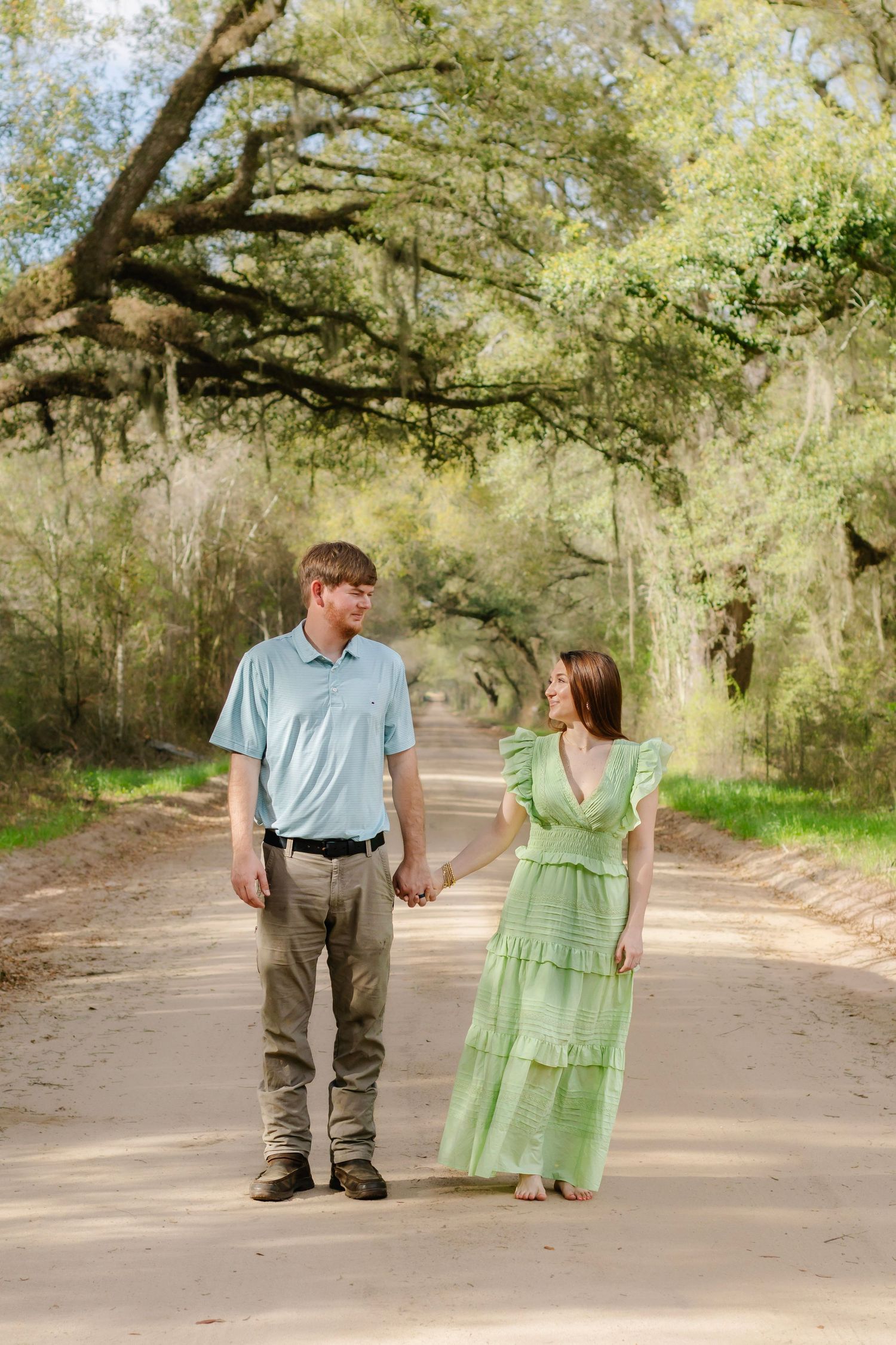 Young couple holding hands on a scenic tree-lined dirt road, woman in green dress, man in blue polo.
