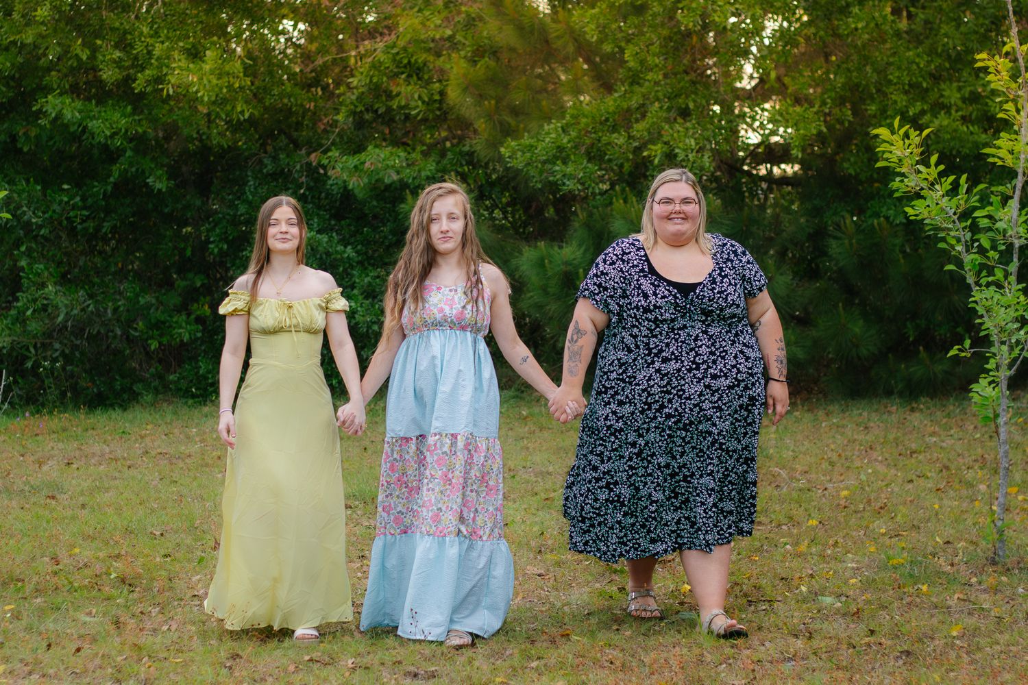 Three females holding hands walking in a green park, wearing colorful dresses outdoors.