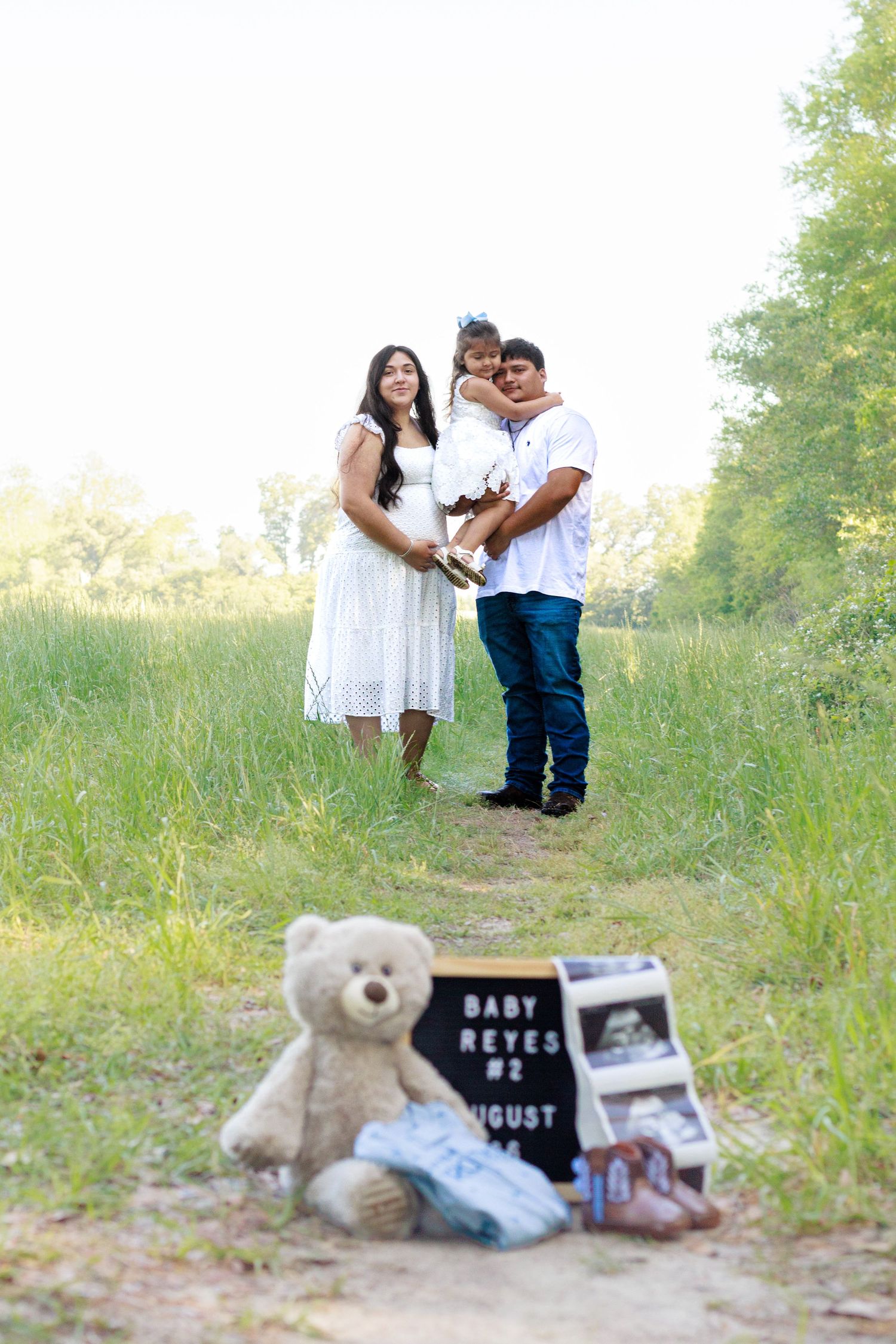 Family pregnancy announcement photo with teddy bear, ultrasound, and Baby Reyes #2 sign in a field.