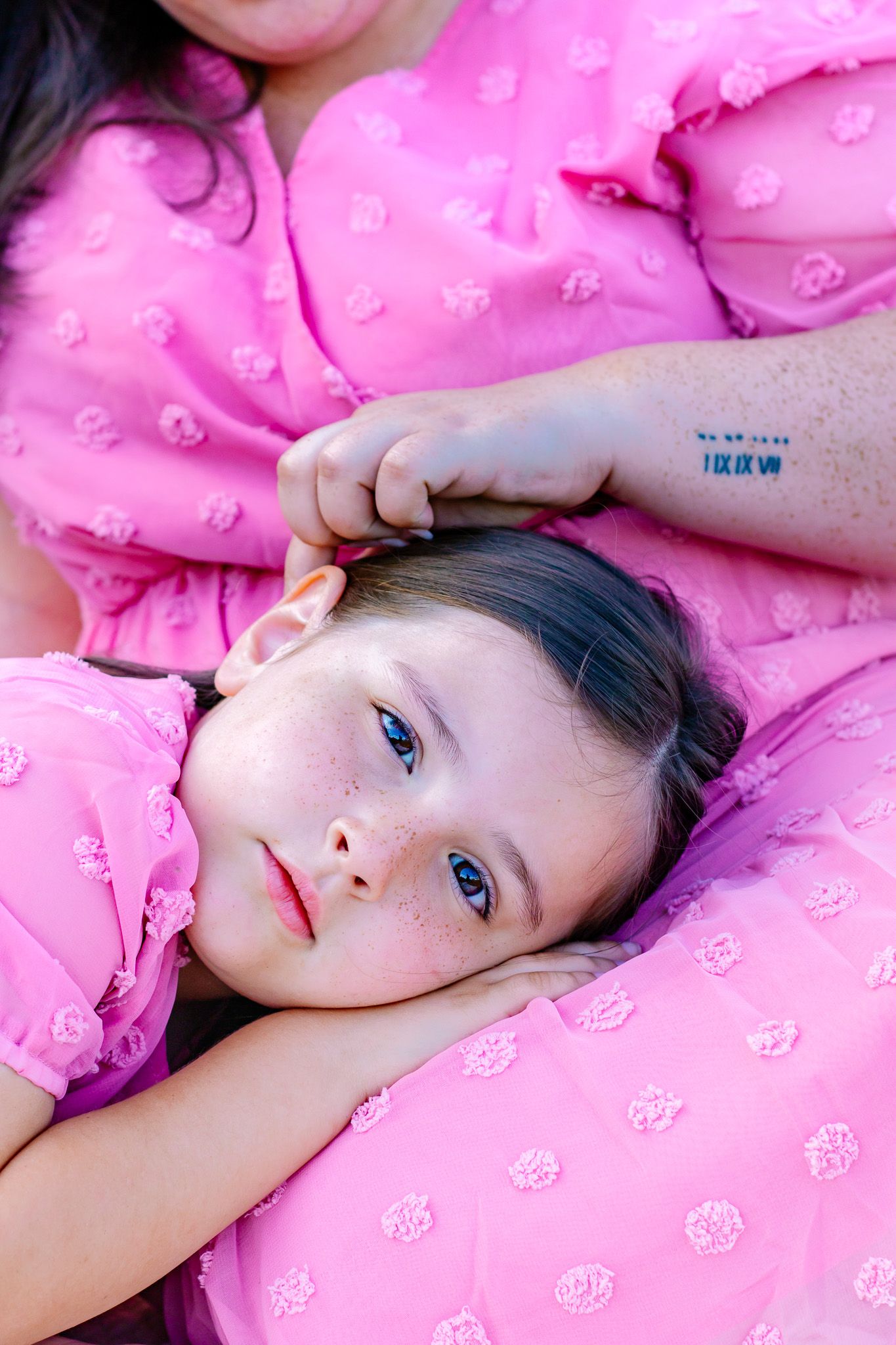 A young girl with blue eyes and freckles rests her head on a pink pillow, wearing a matching pink dress.