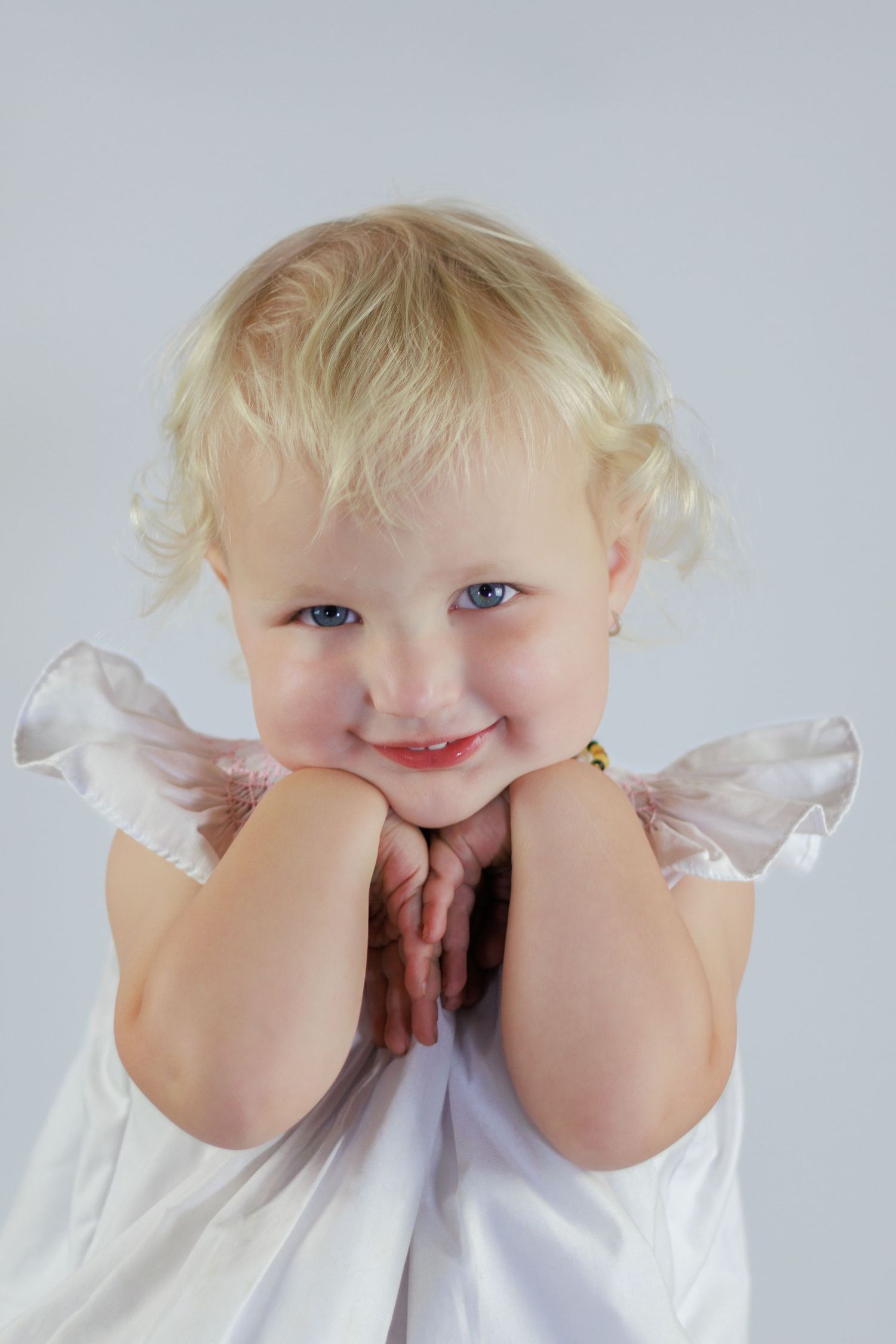 Smiling blonde toddler girl with blue eyes resting chin on hands, wearing white angel dress with wings.