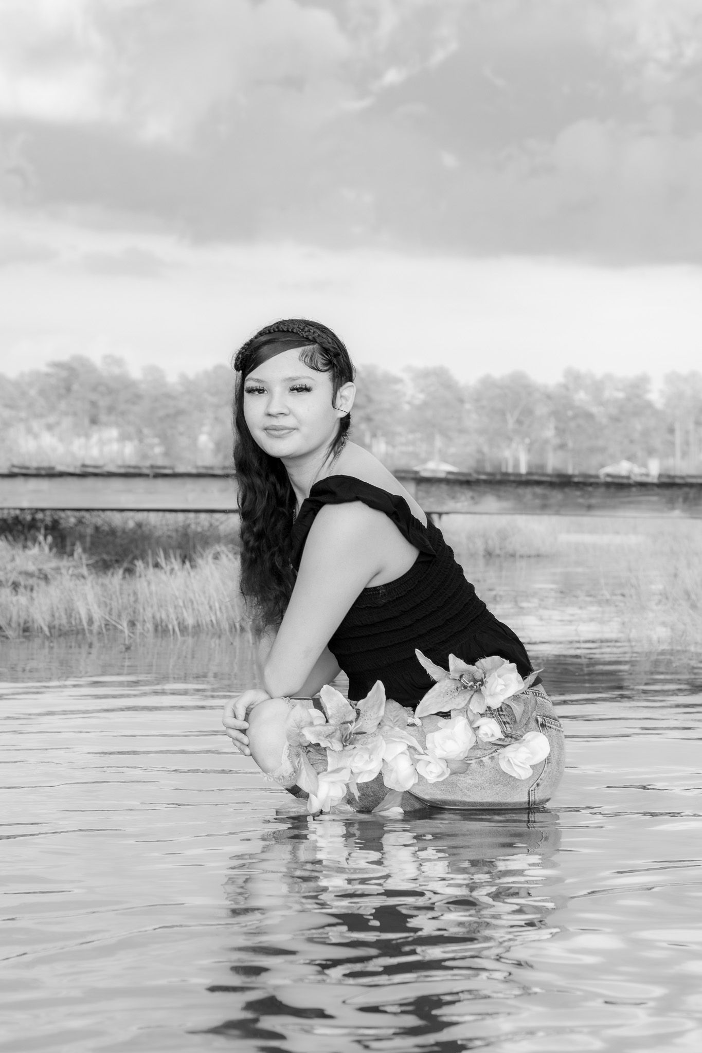 Young woman in black top holds floral wreath while standing in water, black and white portrait photo.