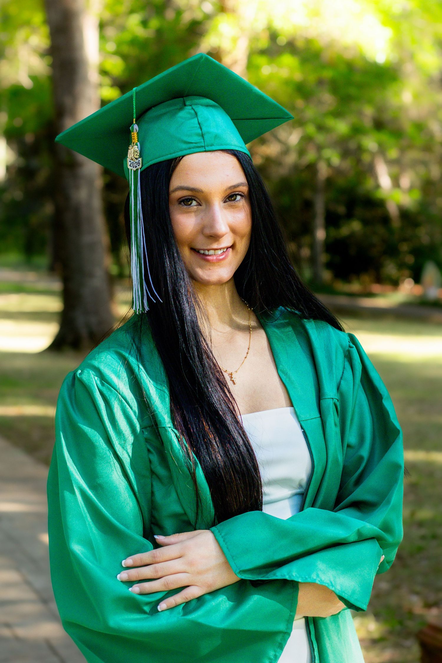Smiling graduate in green cap and gown with arms crossed outdoors.