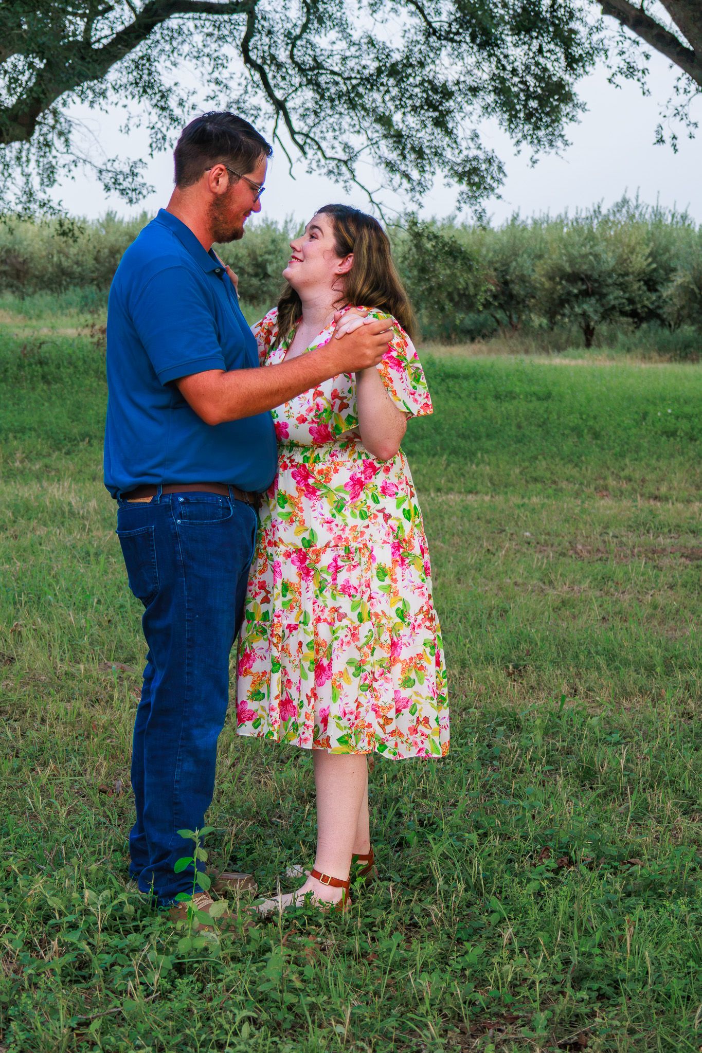 Couple sharing a romantic moment outdoors, woman in floral dress and man in blue shirt embracing in a green field.