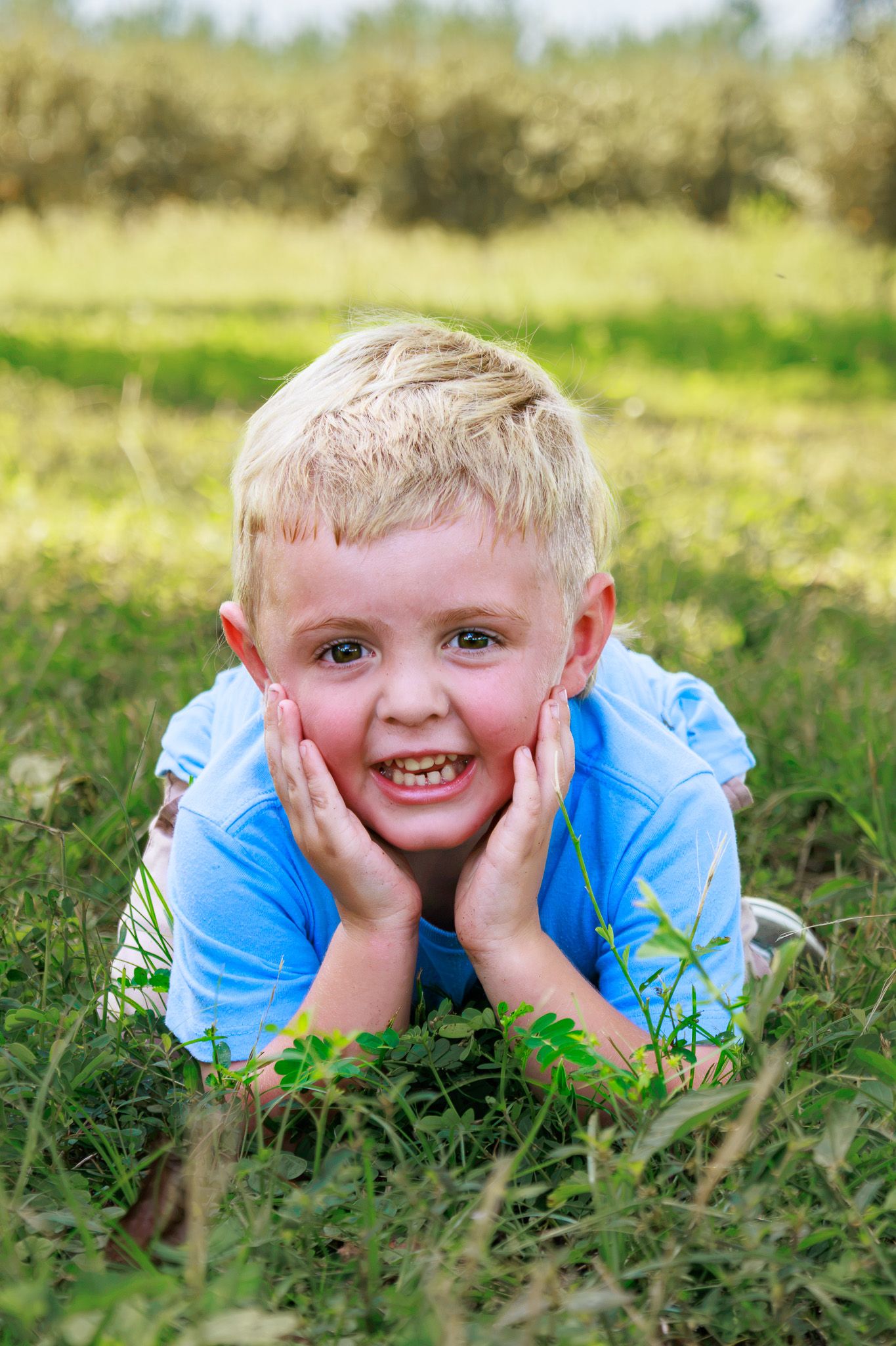 Cheerful blond boy lying on green grass, resting chin in hands, smiling outdoors on a sunny day.