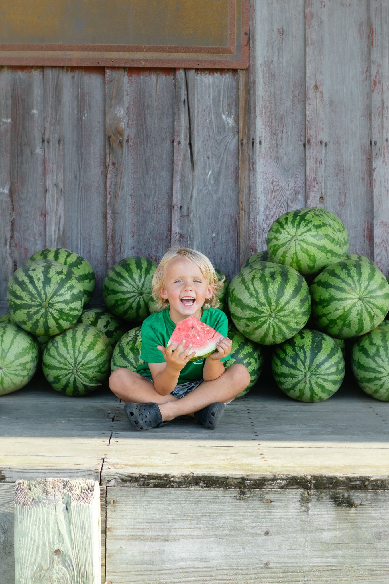 Laughing blonde boy in green shirt sitting cross-legged with watermelon slice, surrounded by stacked watermelons on rustic wooden porch.