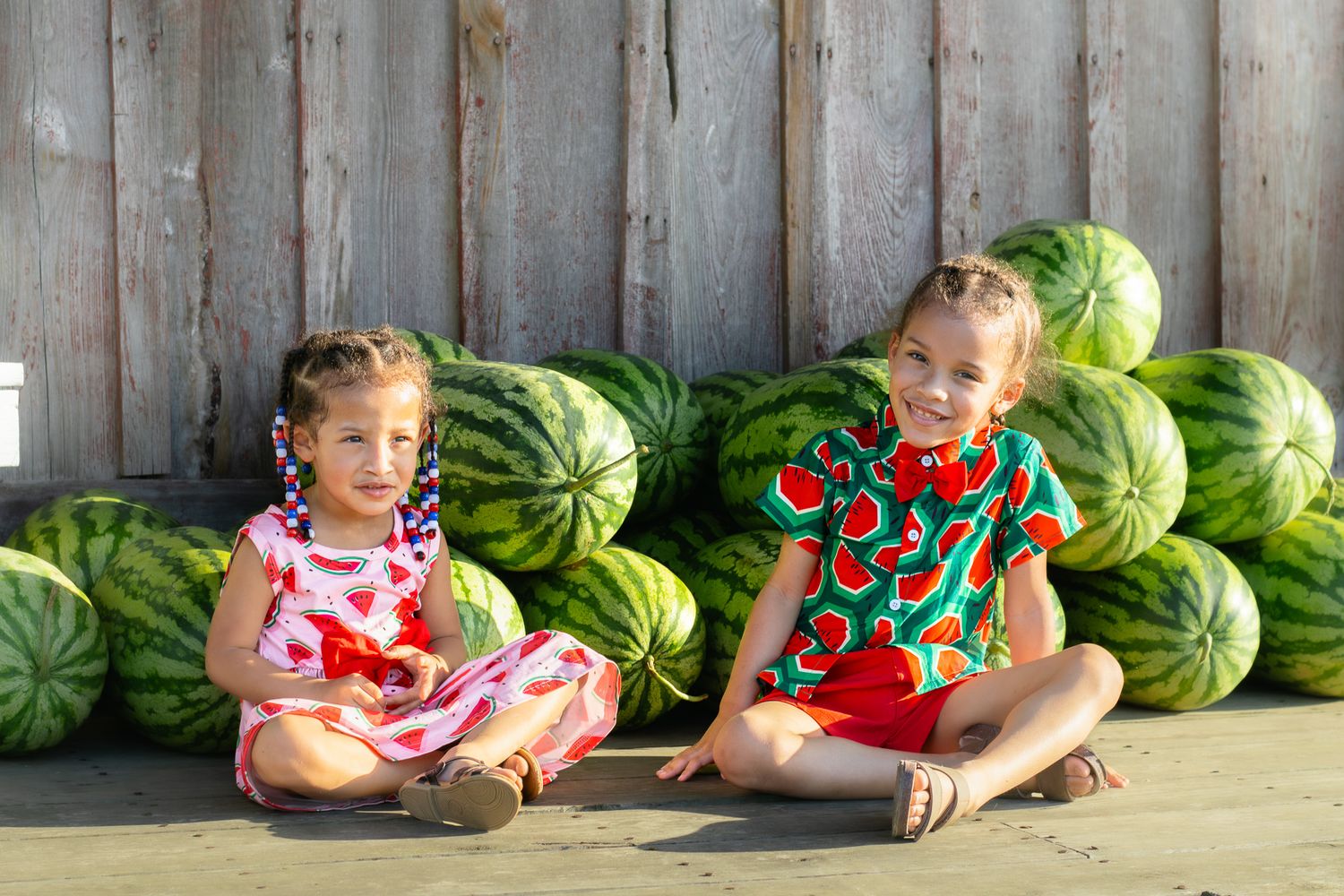 Two young girls wearing watermelon-themed clothing sit together beside a large stack of watermelons.