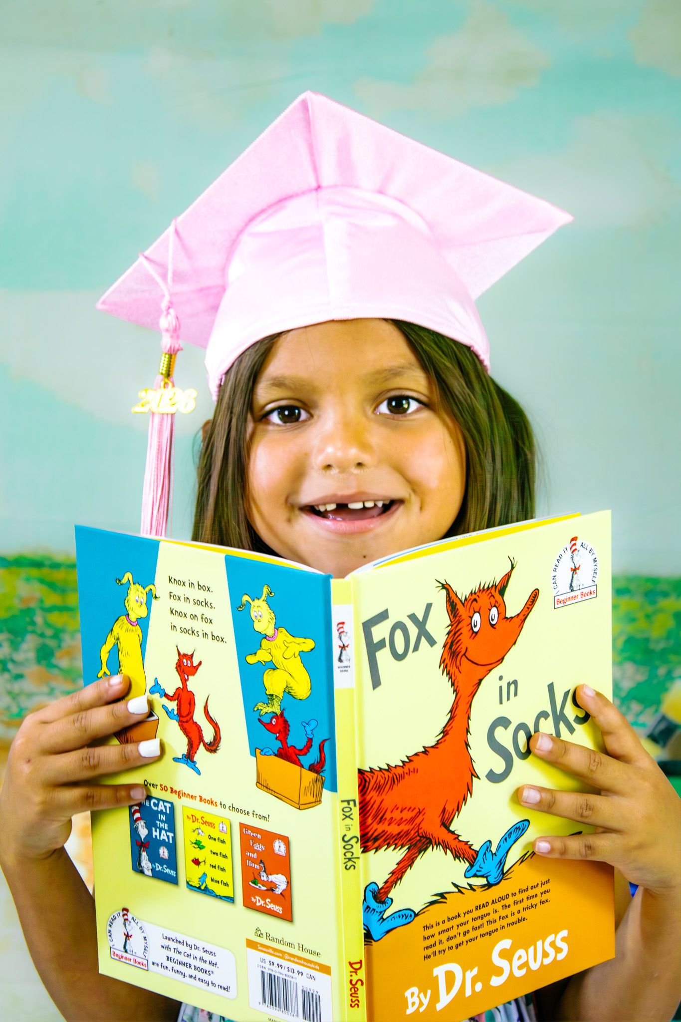 Young girl in pink graduation cap smiling while reading Fox in Socks by Dr. Seuss.