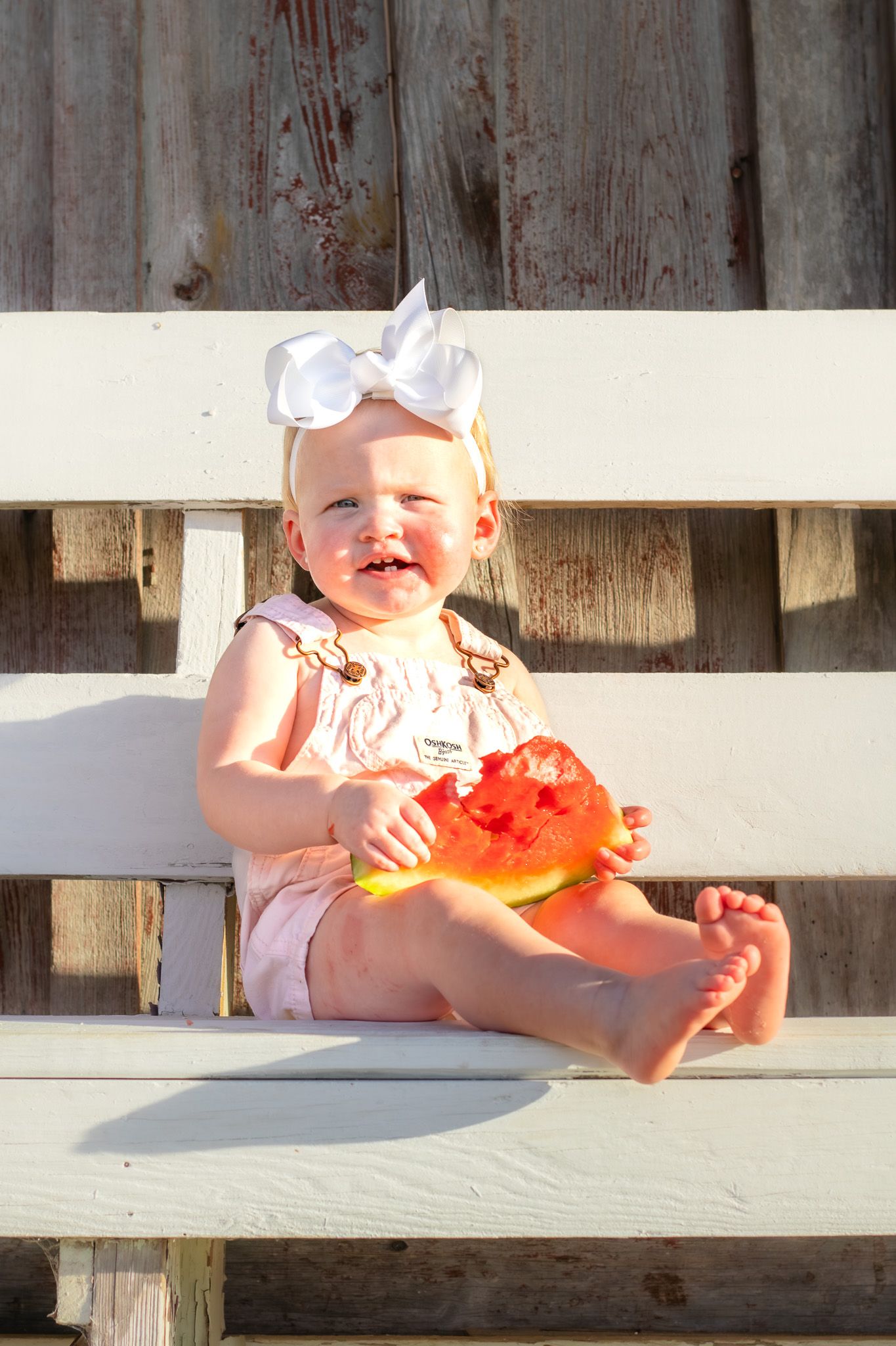 Smiling baby girl with white bow eating watermelon slice on white wooden bench outdoors.