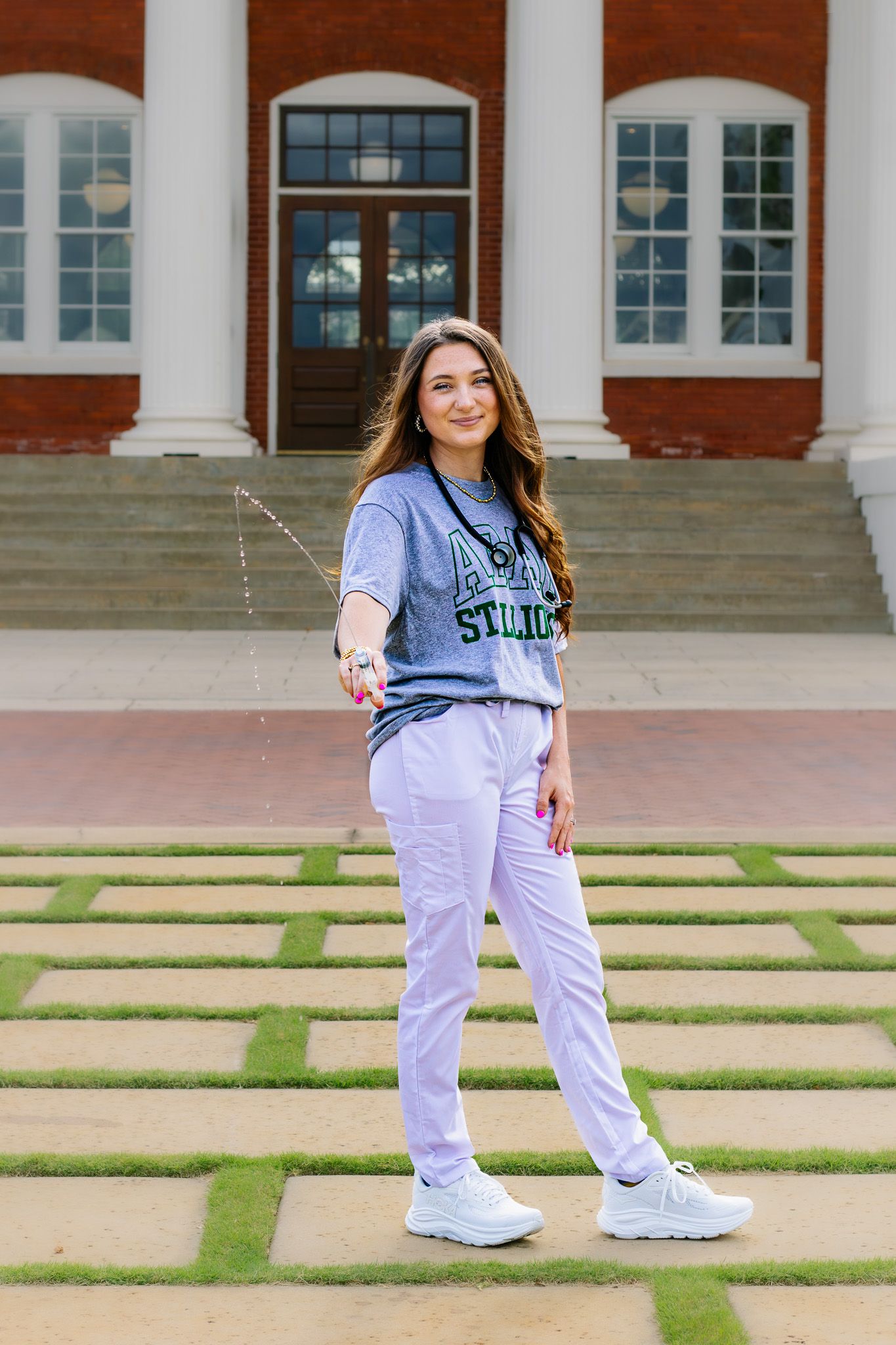 Smiling young woman in college t-shirt and lavender pants posing outside brick university building.
