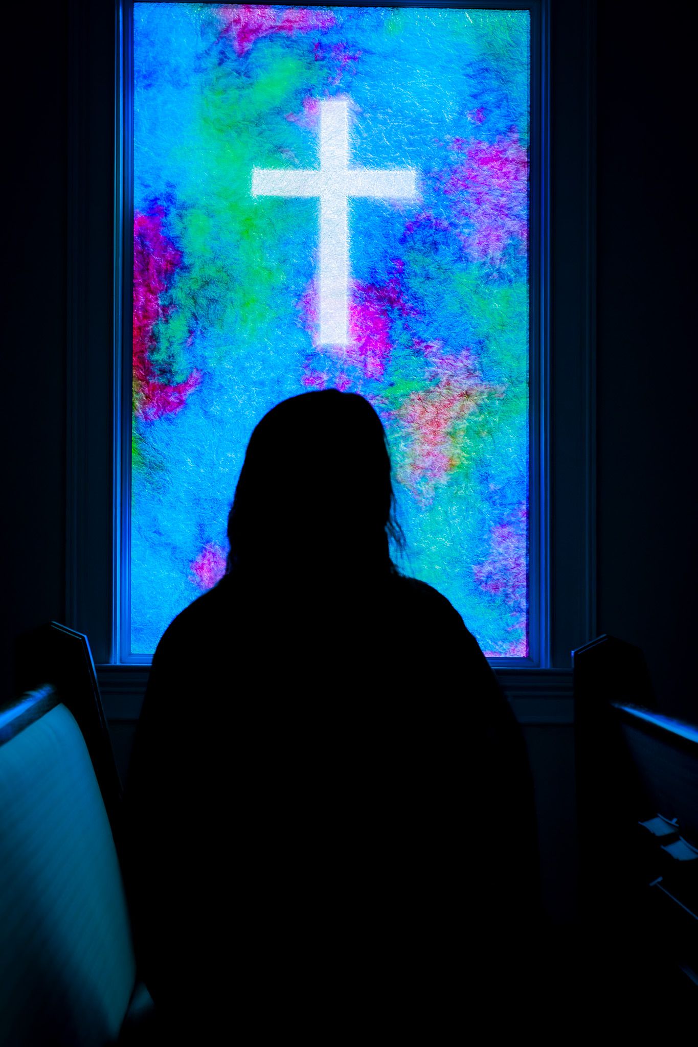 Silhouette of a person sitting before a glowing stained glass cross window in a dark church.