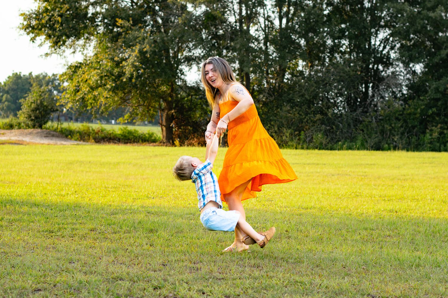 Woman in orange dress joyfully swinging a child in a sunny green park with trees in the background.