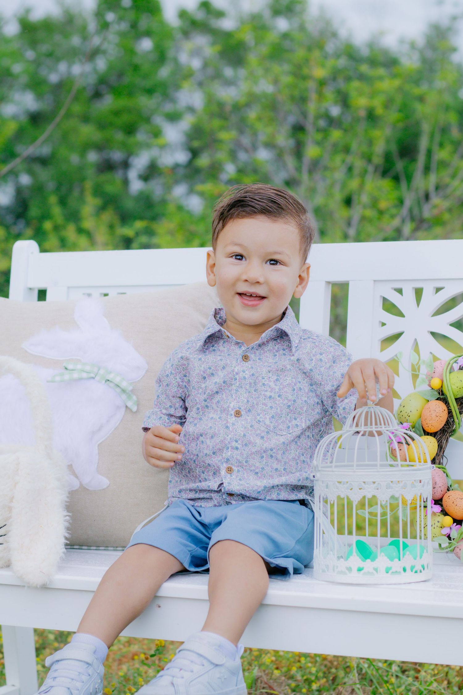 Happy toddler boy sitting on white bench with Easter bunny pillow and decorative birdcage in garden.