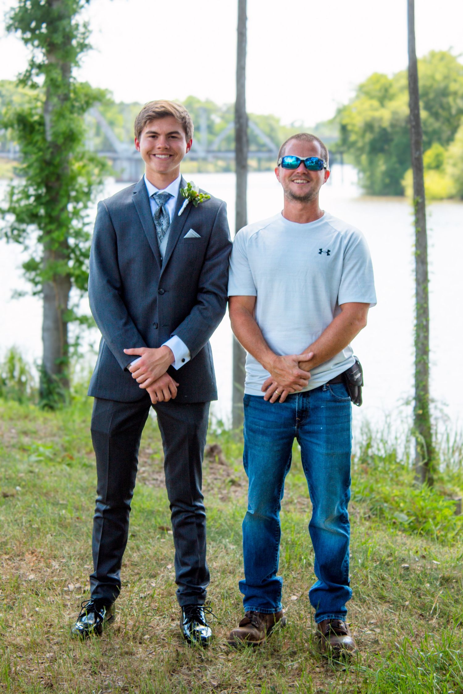 Two men posing outdoors near a river, one in a gray suit with boutonniere and one in casual jeans and white shirt.