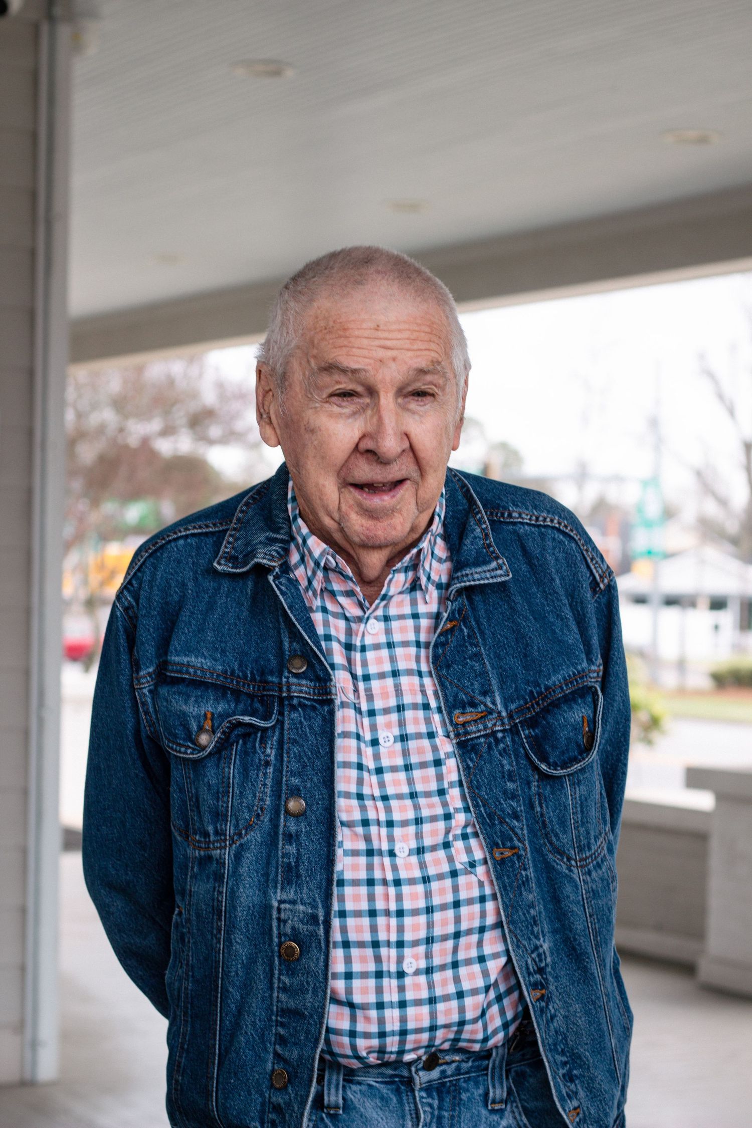 Elderly man in denim jacket and plaid shirt standing outdoors smiling at camera.