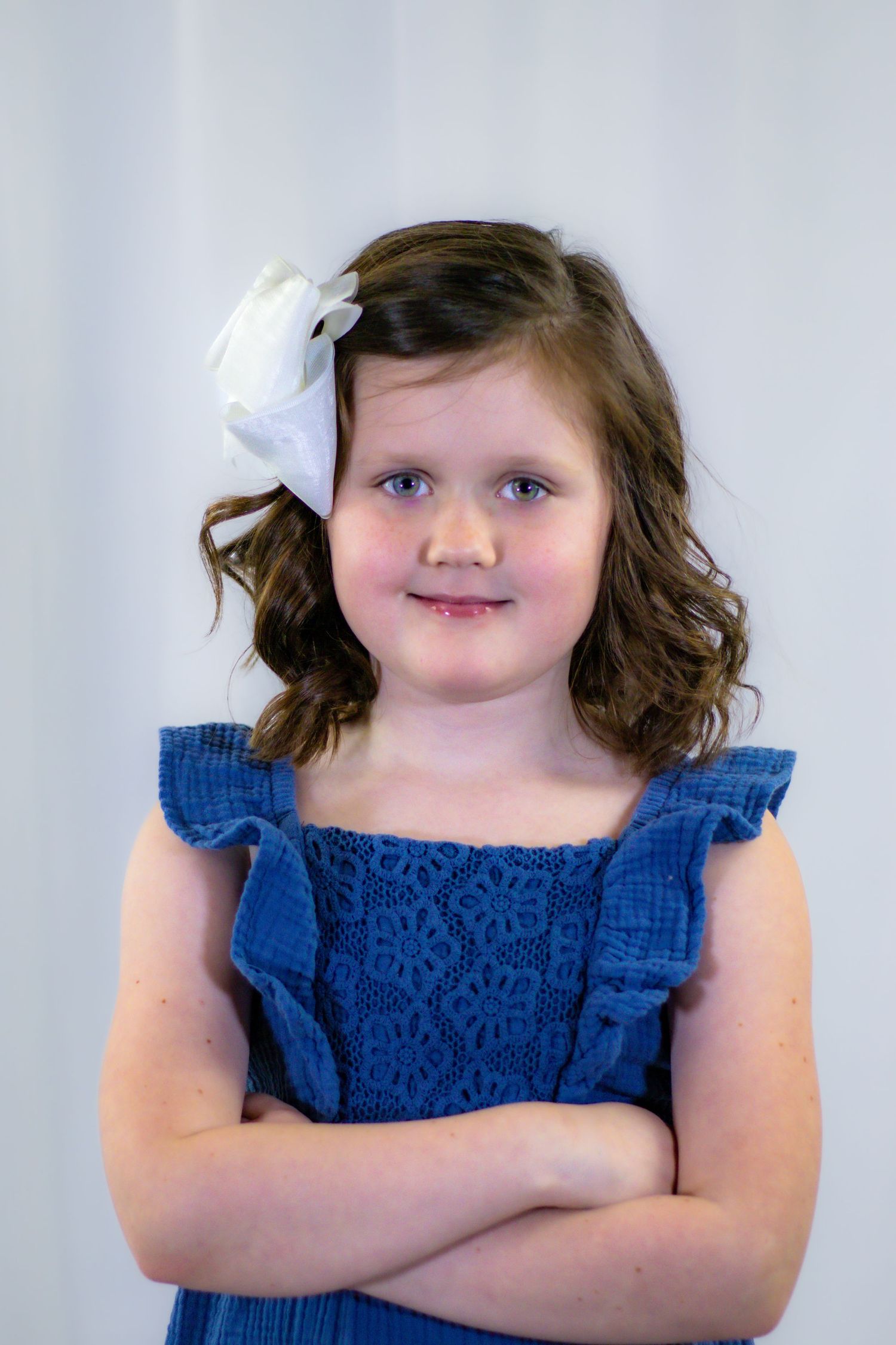 Young girl in blue dress with white bow smiling with arms crossed confidently.
