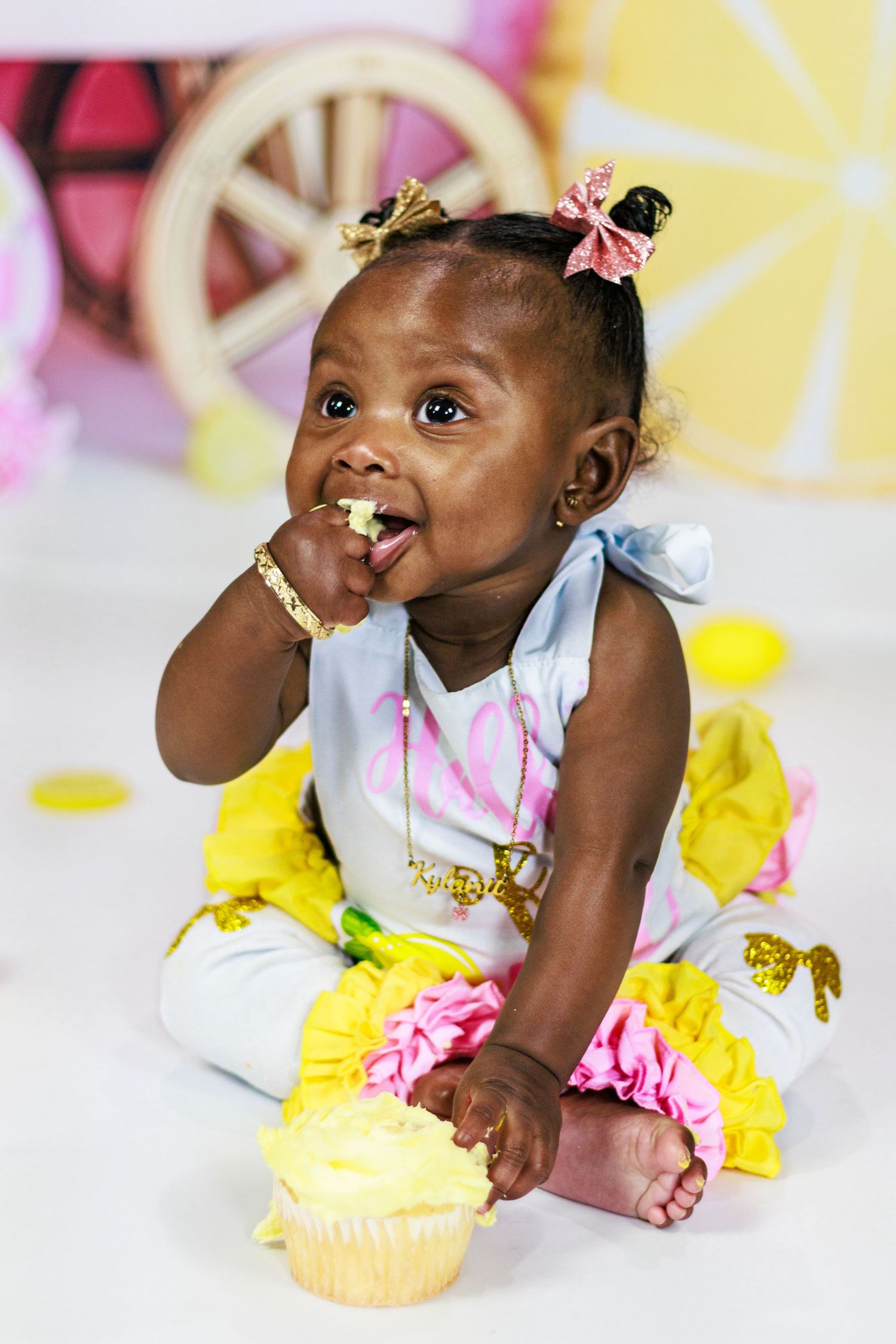 Baby girl with gold crown bows eating a yellow cupcake during a lemon-themed first birthday cake smash session.