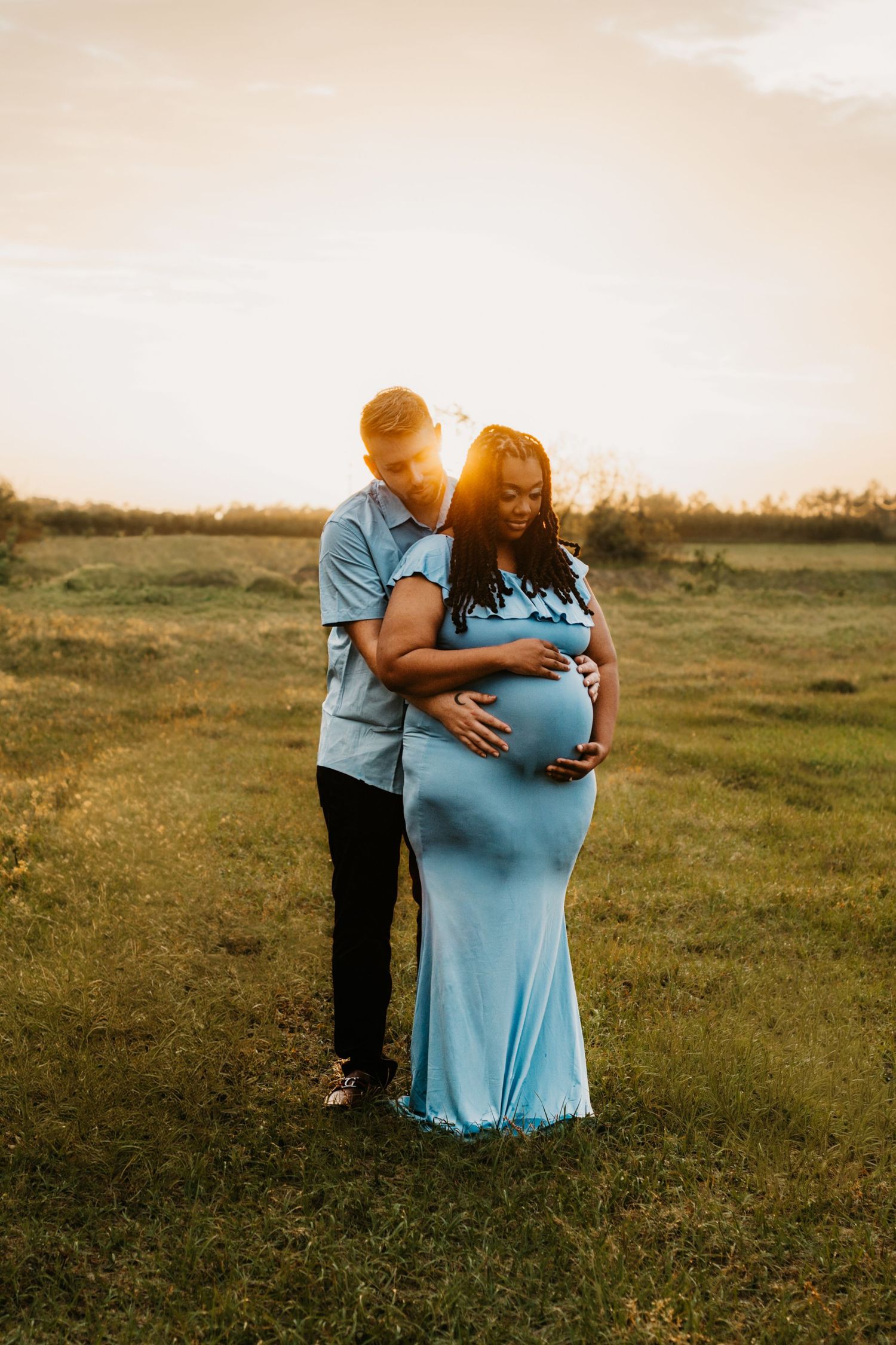 Couple shares intimate moment in glowing sunset field, man embracing pregnant woman in blue gown.