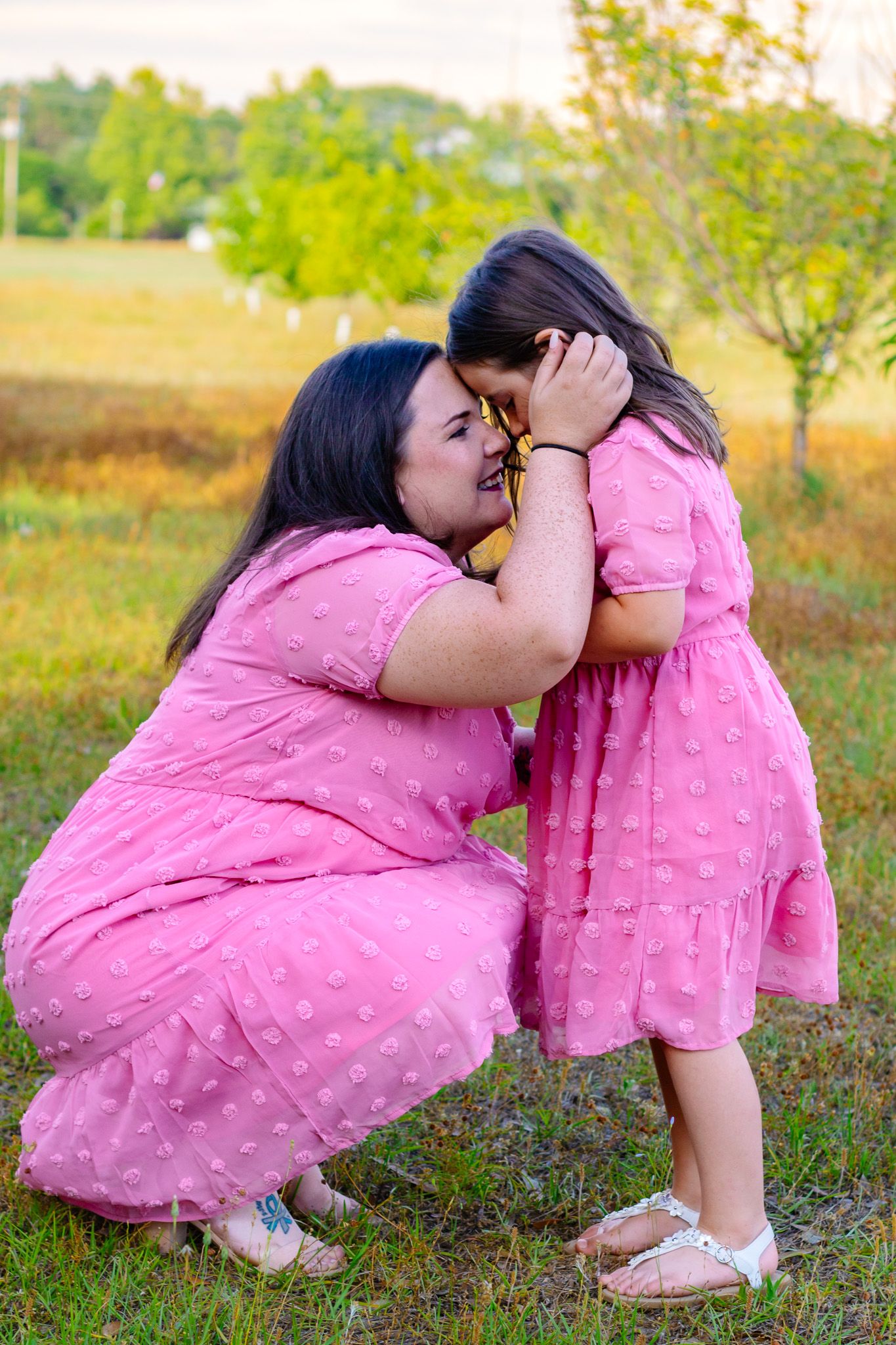 Mother and daughter in matching pink dresses share a tender moment outdoors in a green field.