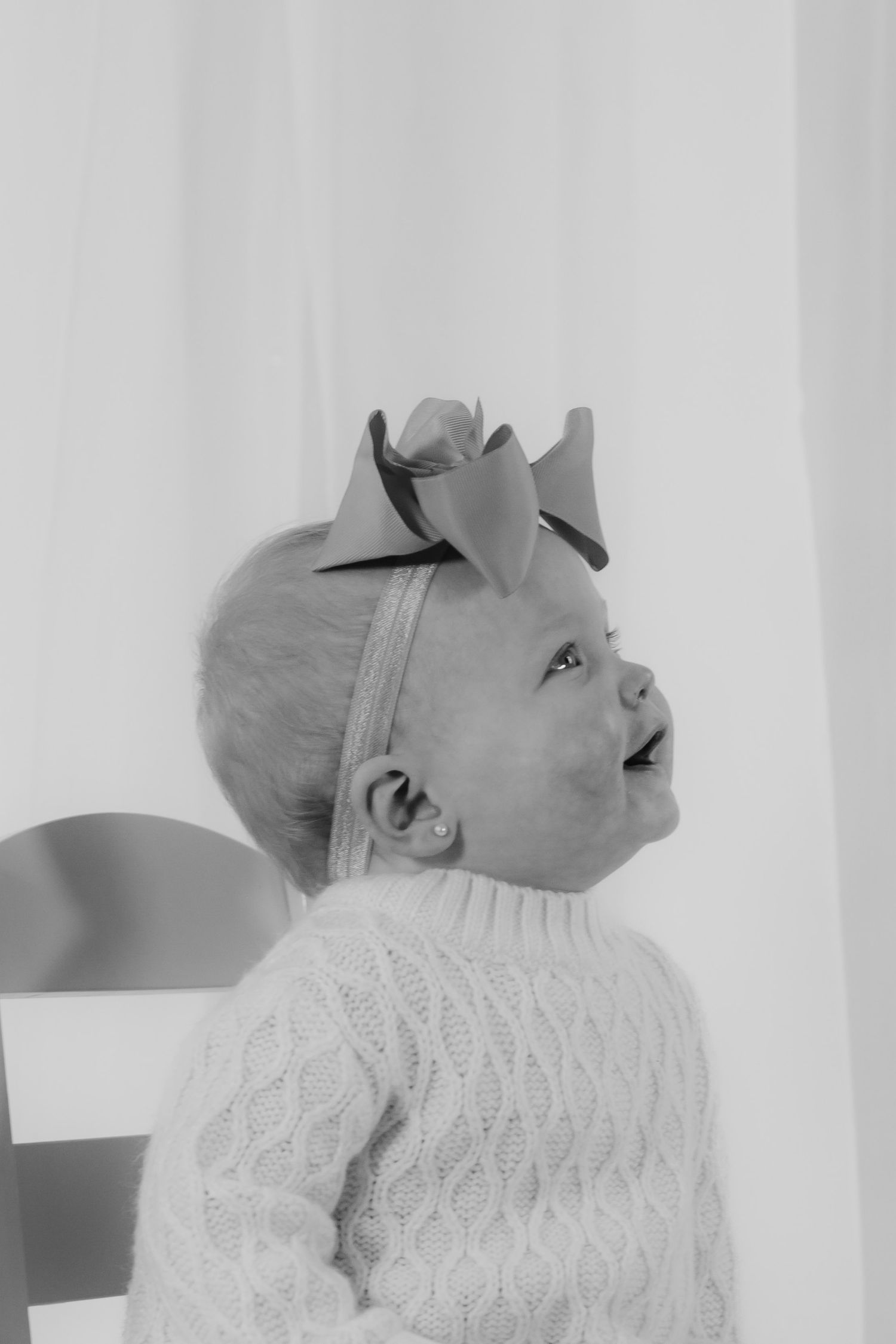 Black and white side profile of a baby girl in cable knit sweater wearing a bow headband looking up.