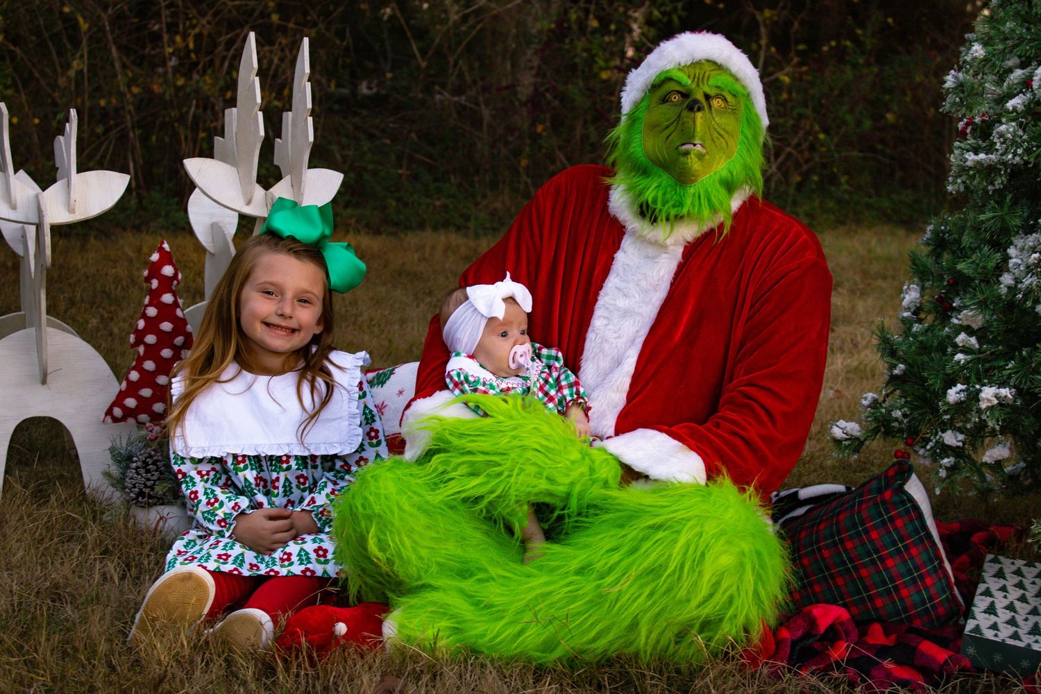 Young girl and infant posing with Grinch character in Santa suit beside Christmas tree outdoors.