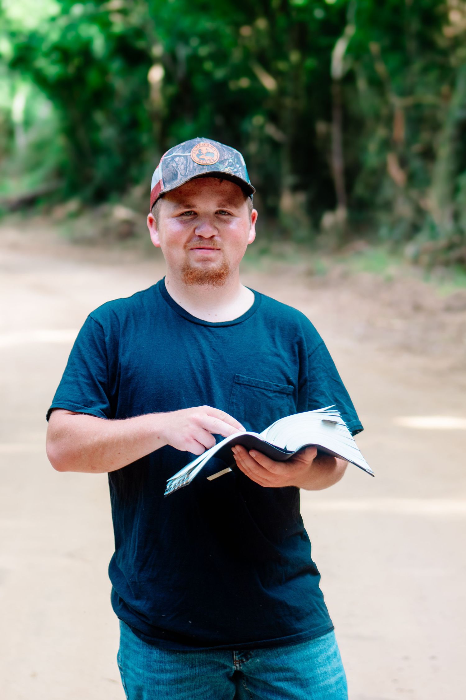 Man in black t-shirt and camo cap holding an open book on a dirt road surrounded by green trees.