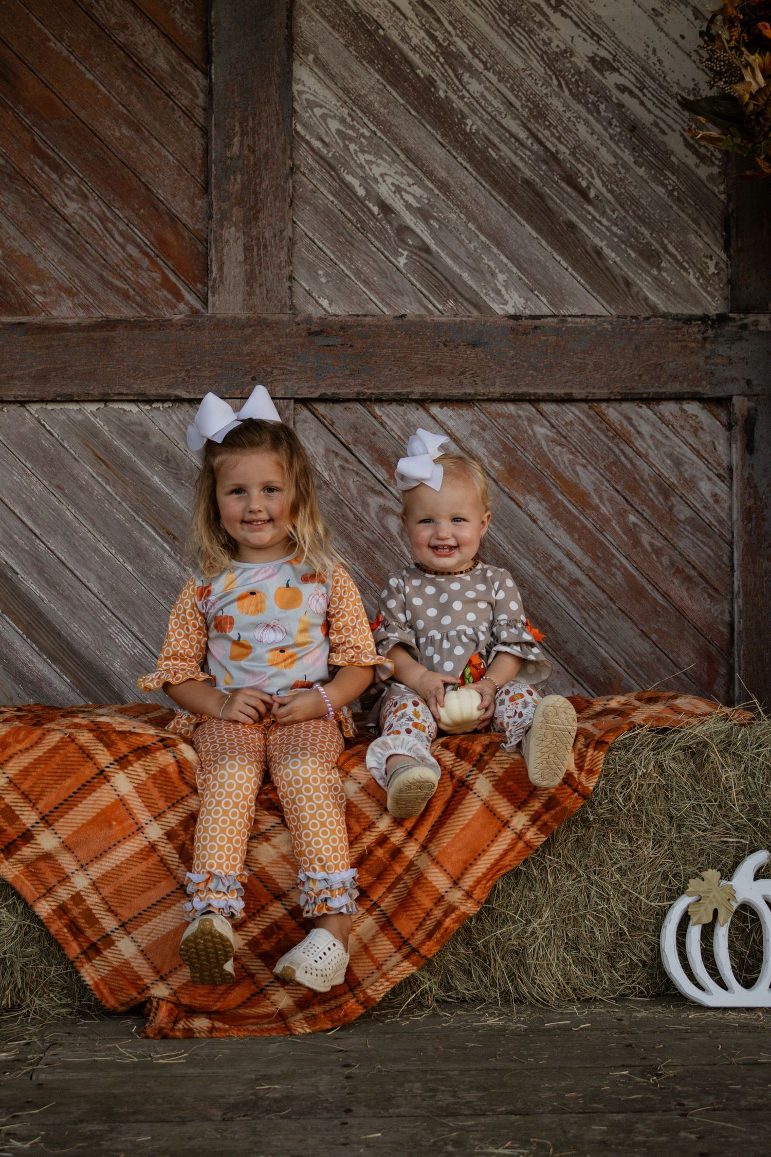 Two little girls in matching fall outfits sitting on hay bale with white bow headbands.