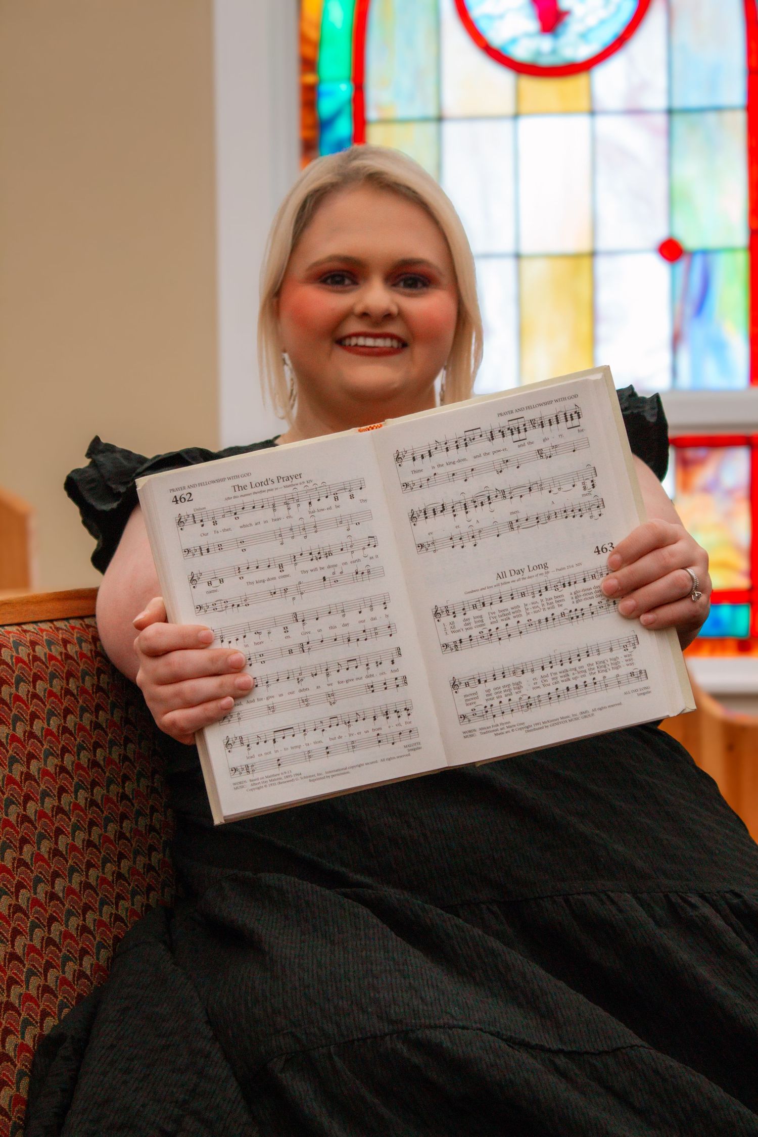 Smiling blonde woman sitting in church pew holding open a hymnal with stained glass windows behind her.