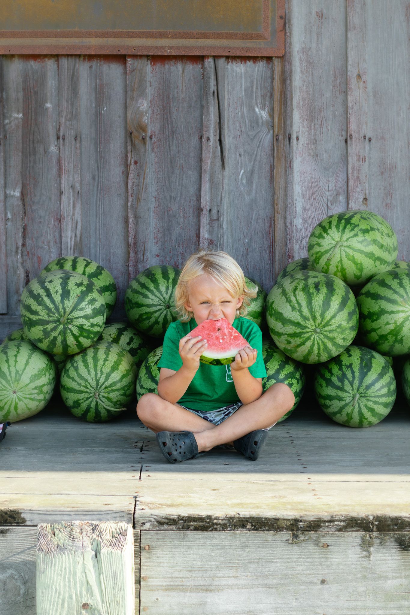 Blond child sitting cross-legged eating a watermelon slice in front of stacked whole watermelons by a rustic wooden wall.