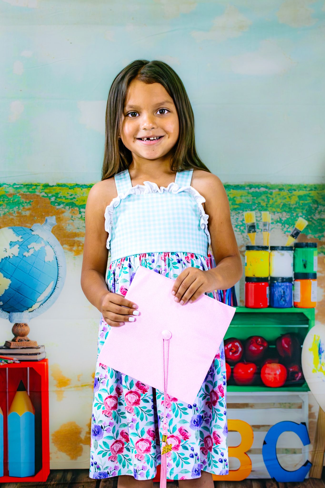 Smiling young girl in floral dress holding pink paper in colorful classroom with globe and art supplies.