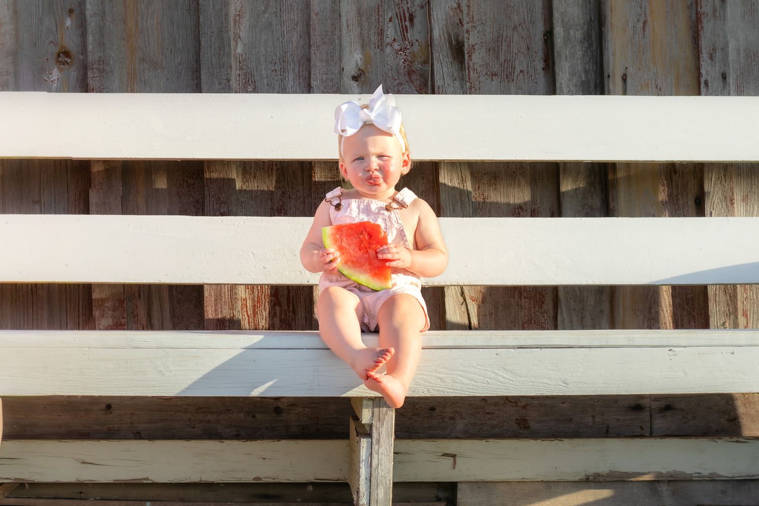 Happy baby girl in pink outfit sitting on wooden fence holding a large slice of watermelon.