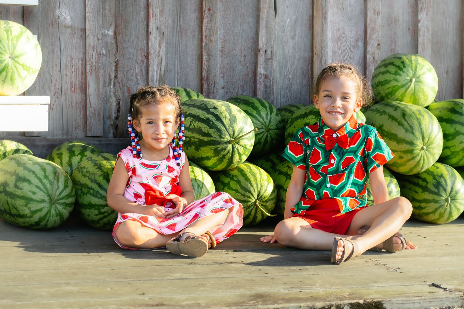 Two girls in watermelon-print outfits sit cross-legged in front of a large pile of watermelons outdoors.