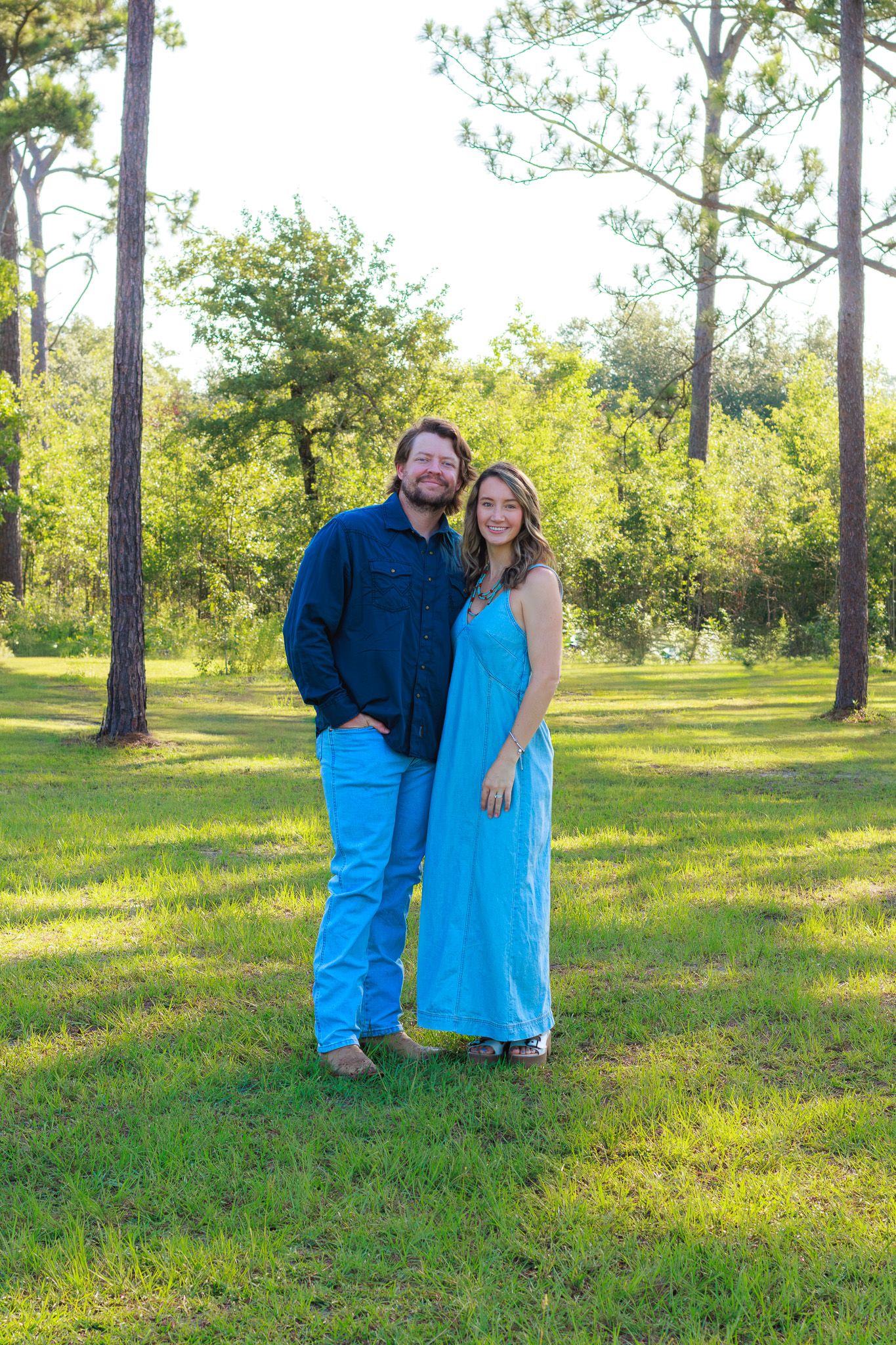 Couple smiling together in a lush green park, man in blue shirt and jeans, woman in turquoise maxi dress.